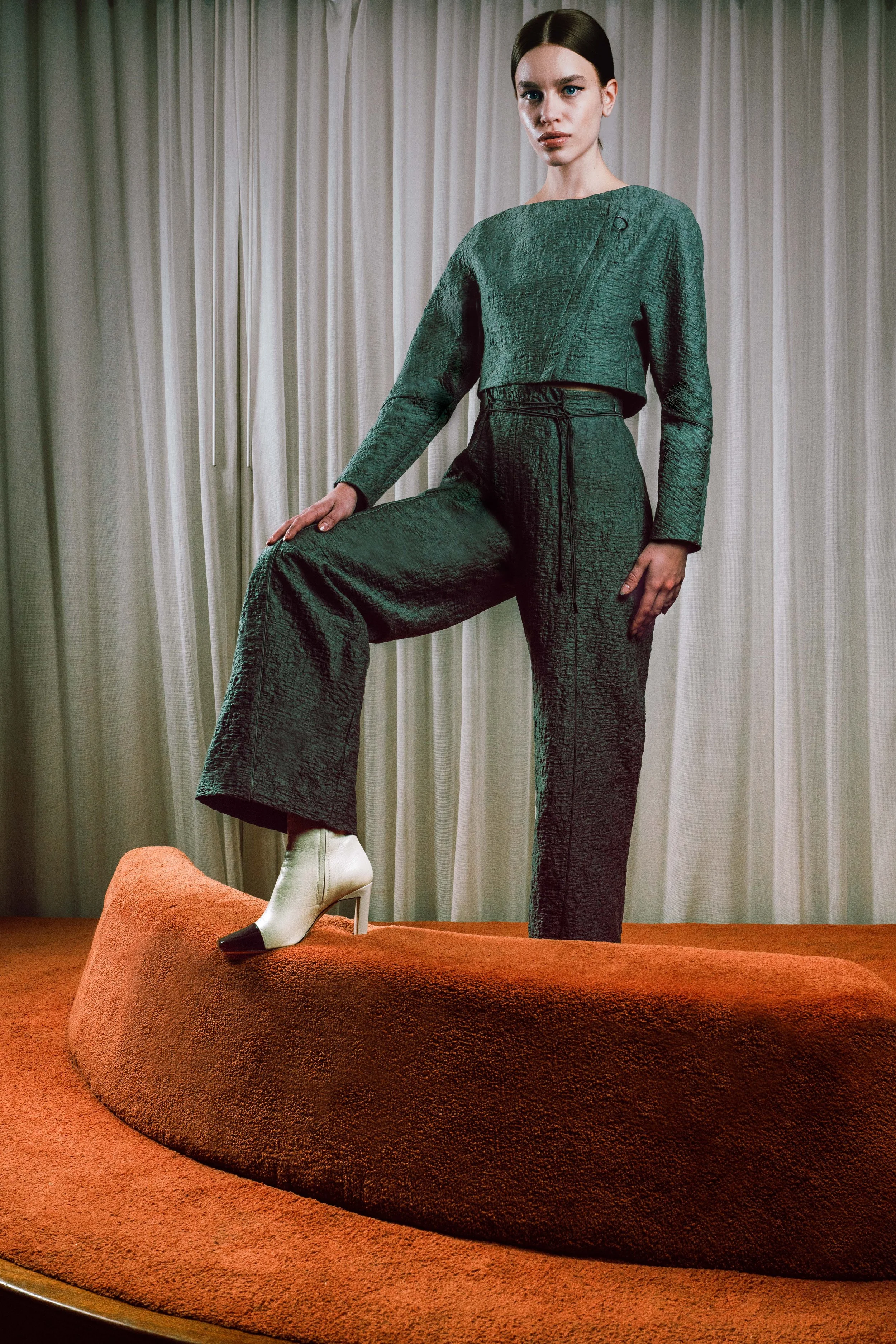 A woman with dark hair in a sleek style, wearing a green textured long-sleeve top and matching pants, standing on a raised orange textured platform with one foot on a chair and the other on the platform, against a background of beige curtains.