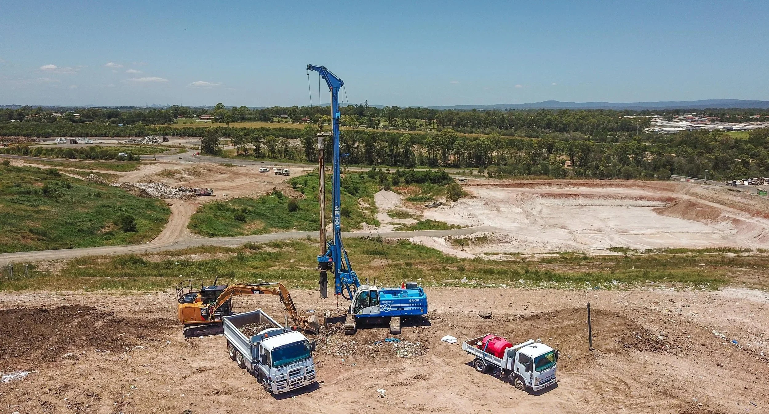 Construction site with heavy machinery including a large blue crane, excavator, and several trucks, with a landscape of fields, trees, and distant buildings under a clear blue sky.