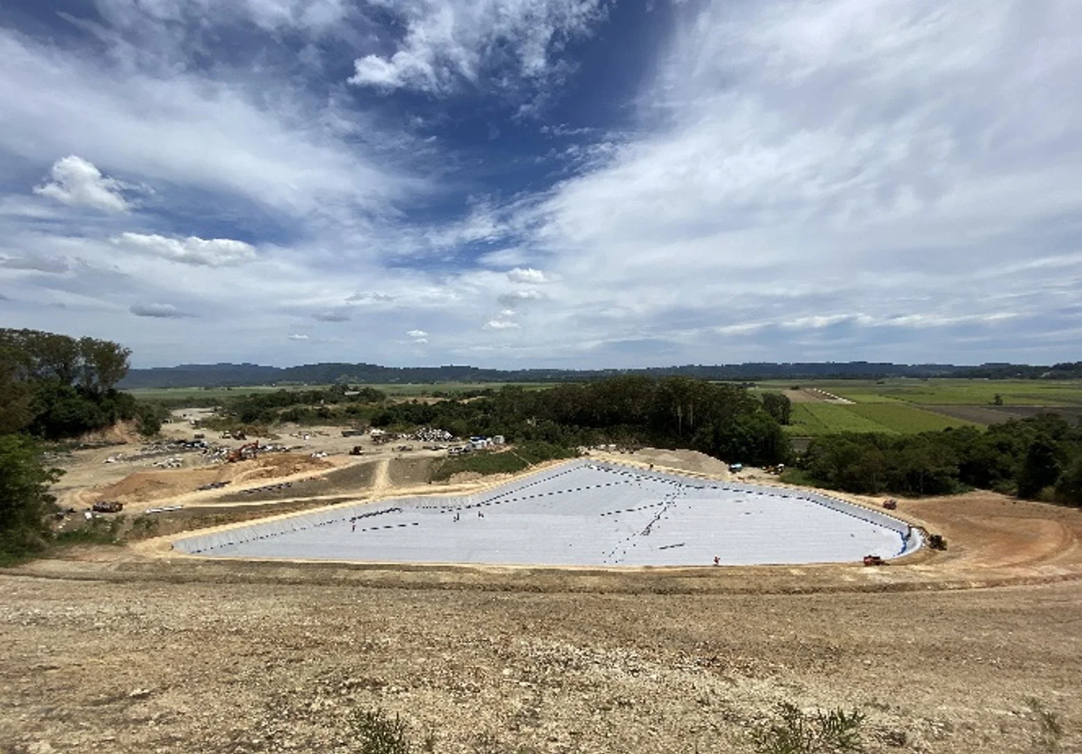 A construction site in a rural area with a large, oval-shaped foundation covered with a white tarp, surrounded by dirt roads and construction vehicles, with fields and a cloudy sky in the background.