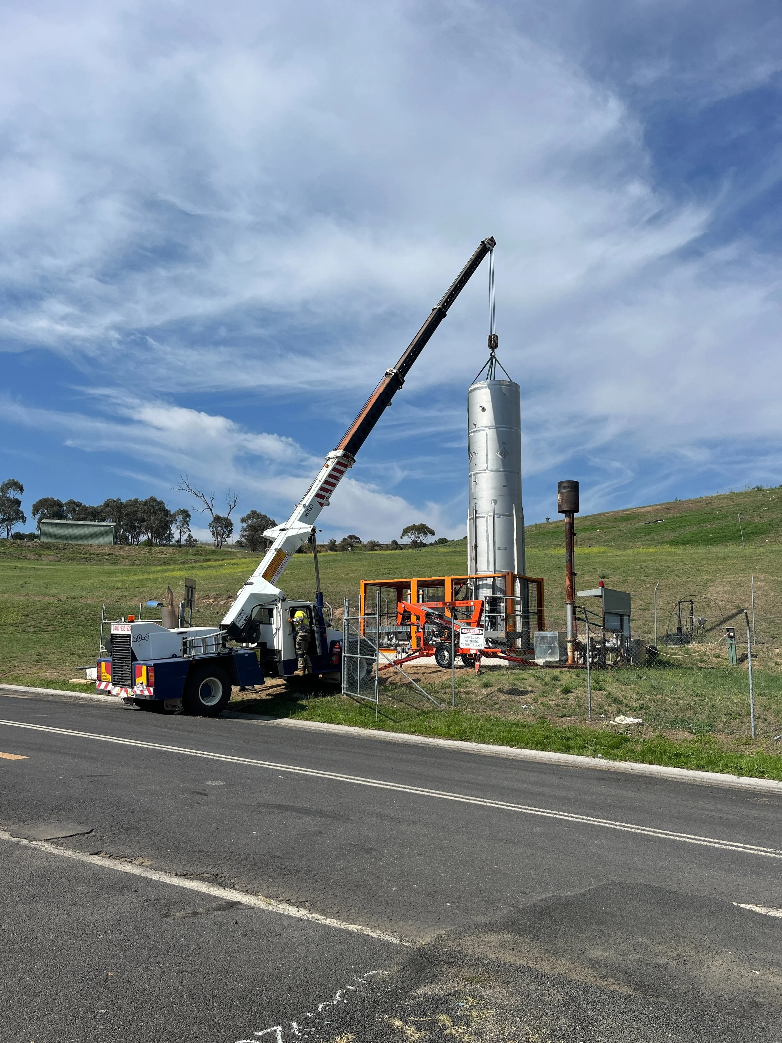 A crane lifting a large cylindrical metal tank on a construction site with a fence, grassy hillside, and a partly cloudy sky in the background.