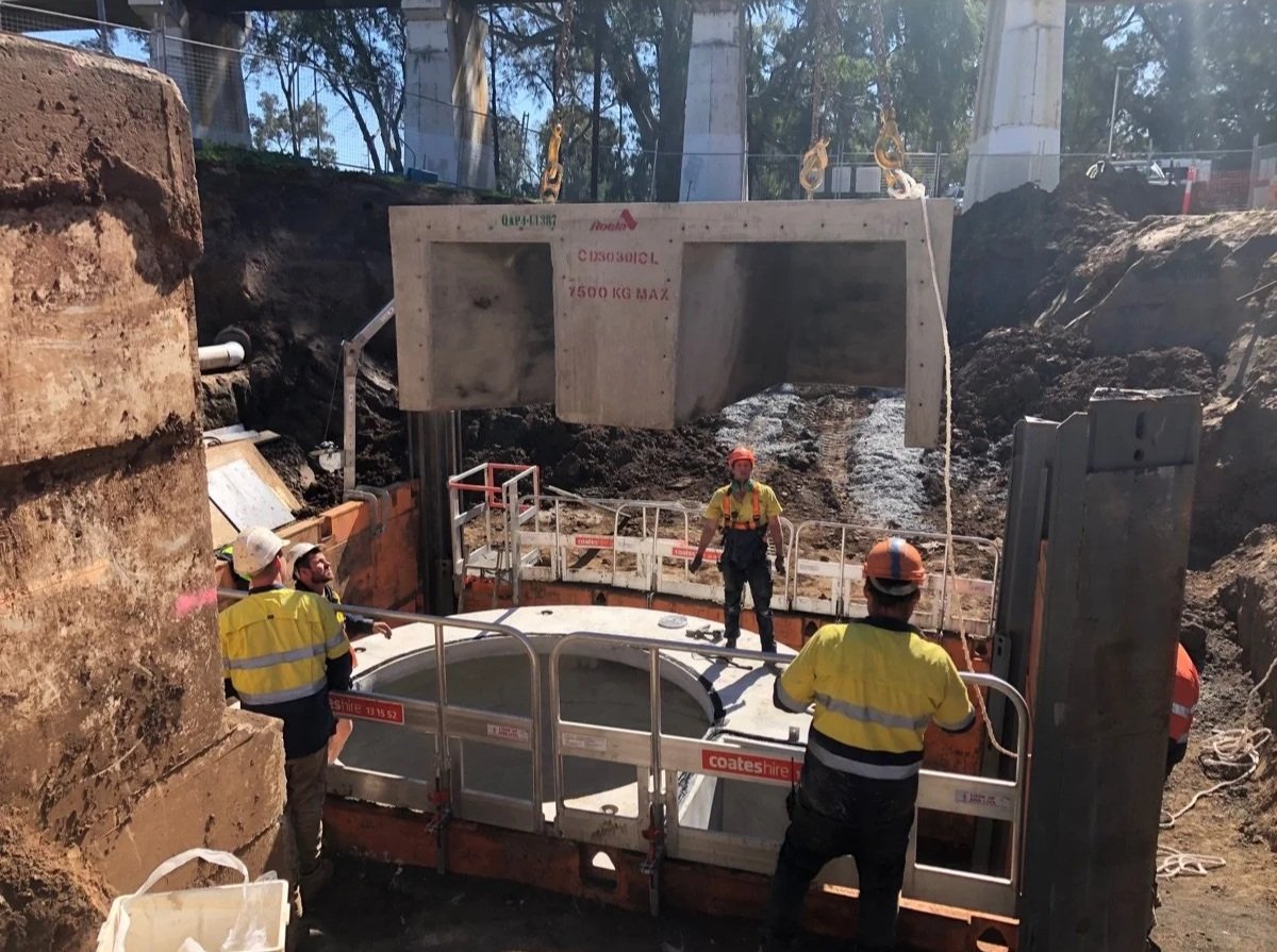 Workers in safety gear operating heavy machinery at an excavation site with large concrete structure being lowered into the ground.