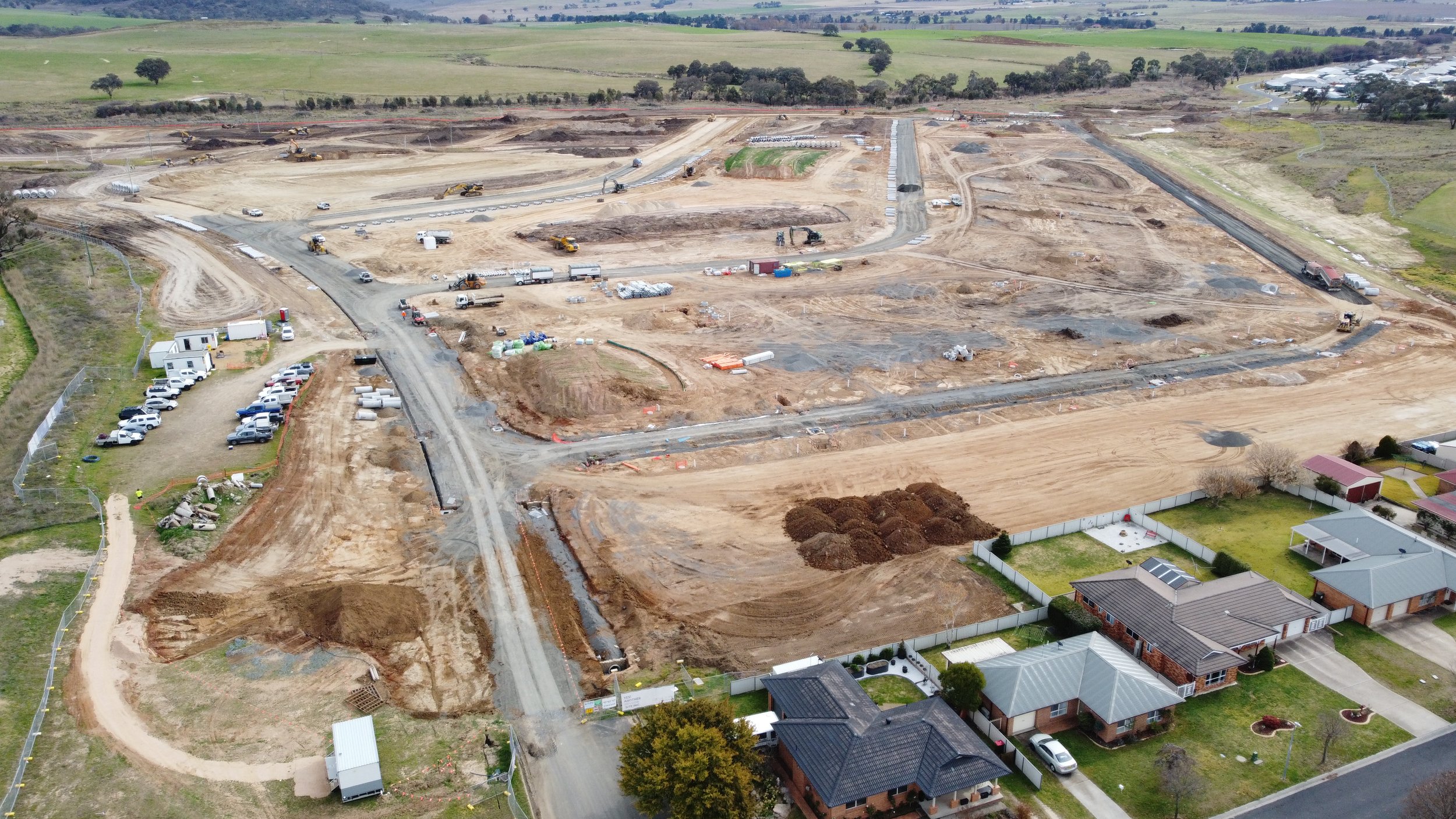 Aerial view of a construction site with dirt roads, heavy machinery, and scattered building materials, surrounded by residential houses and green fields.