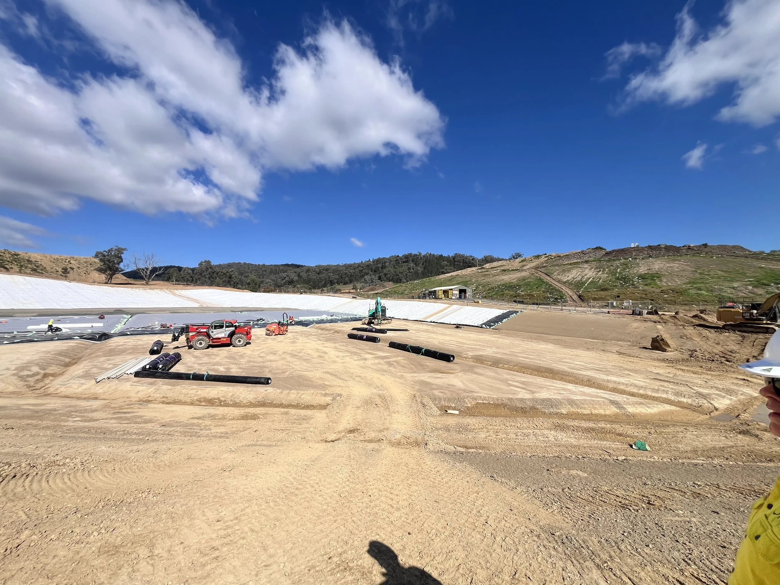 Construction site with large white covers, construction equipment, and workers under a partly cloudy sky with hills in the background.
