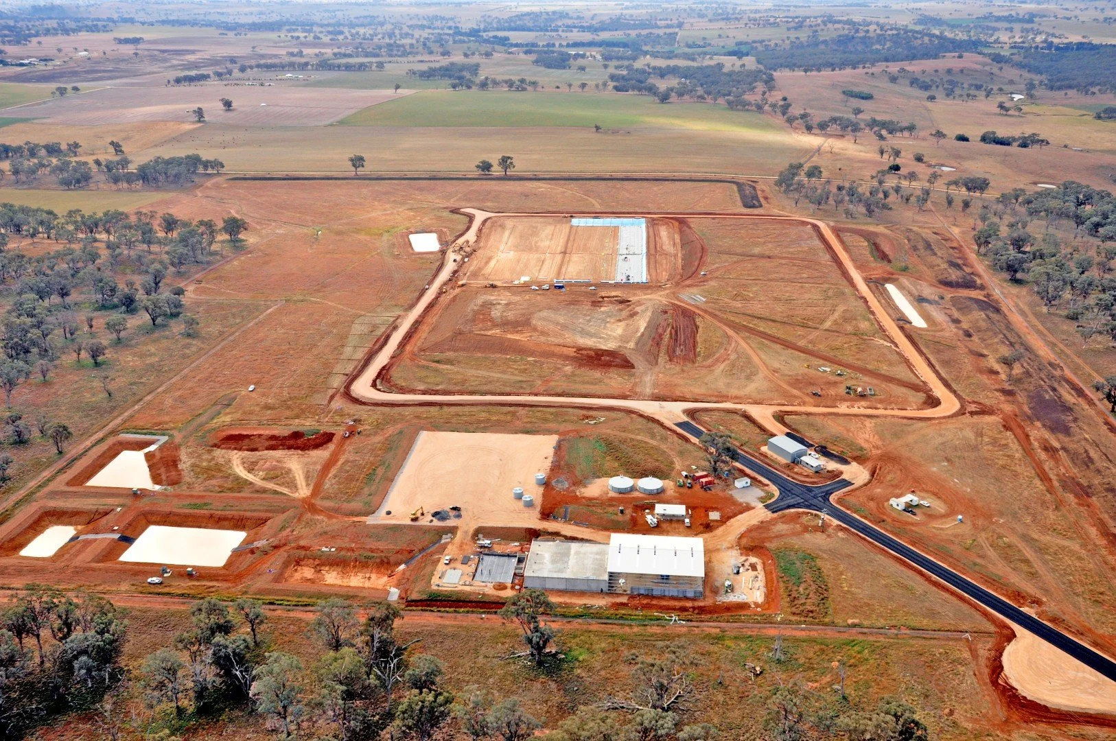 Aerial view of a large construction site in a rural area with dirt roads, several buildings, tanks, and open fields surrounded by trees.