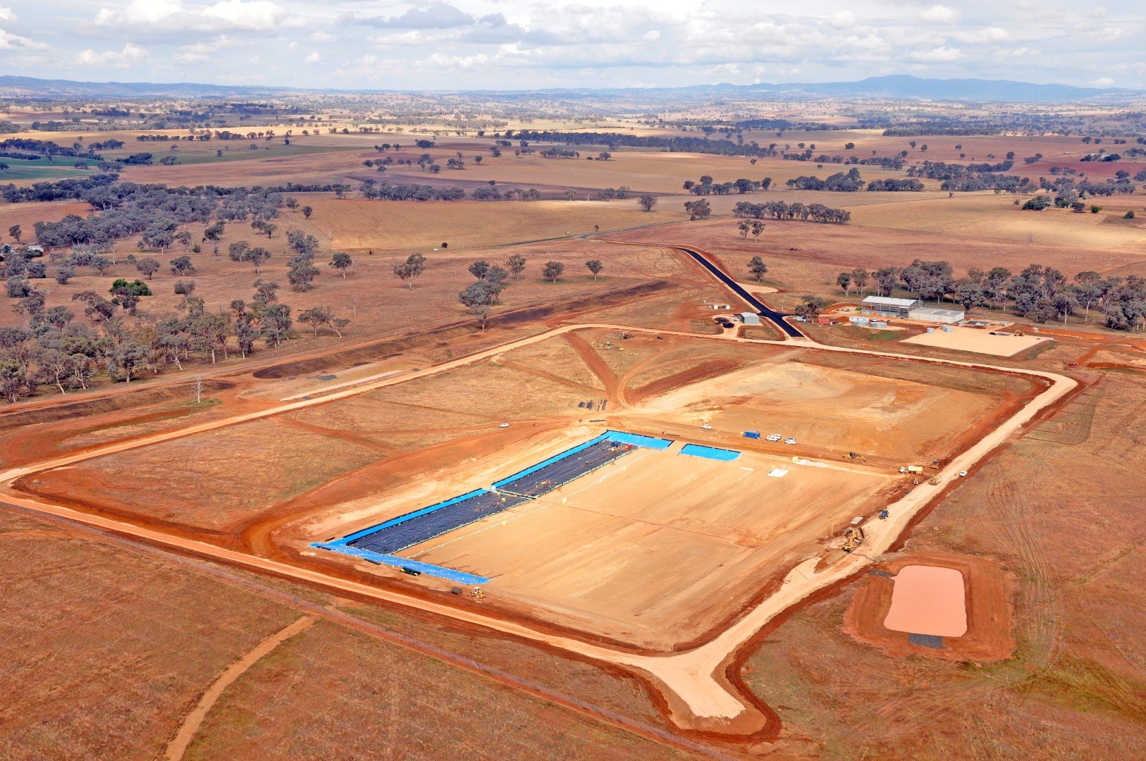 Aerial view of a construction site in a rural area with open fields, dirt roads, and some buildings in the distance. There is a large rectangular area with a black surface and a small pond nearby.