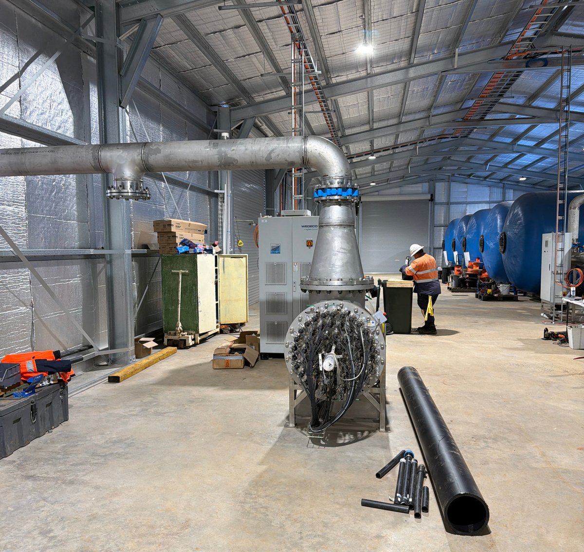Industrial warehouse with large metal piping, a worker in safety gear, and blue water tanks in the background.