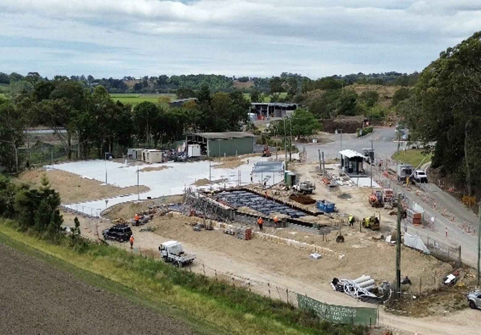 Construction site with workers, machinery, and a partially built road, surrounded by trees and greenery.