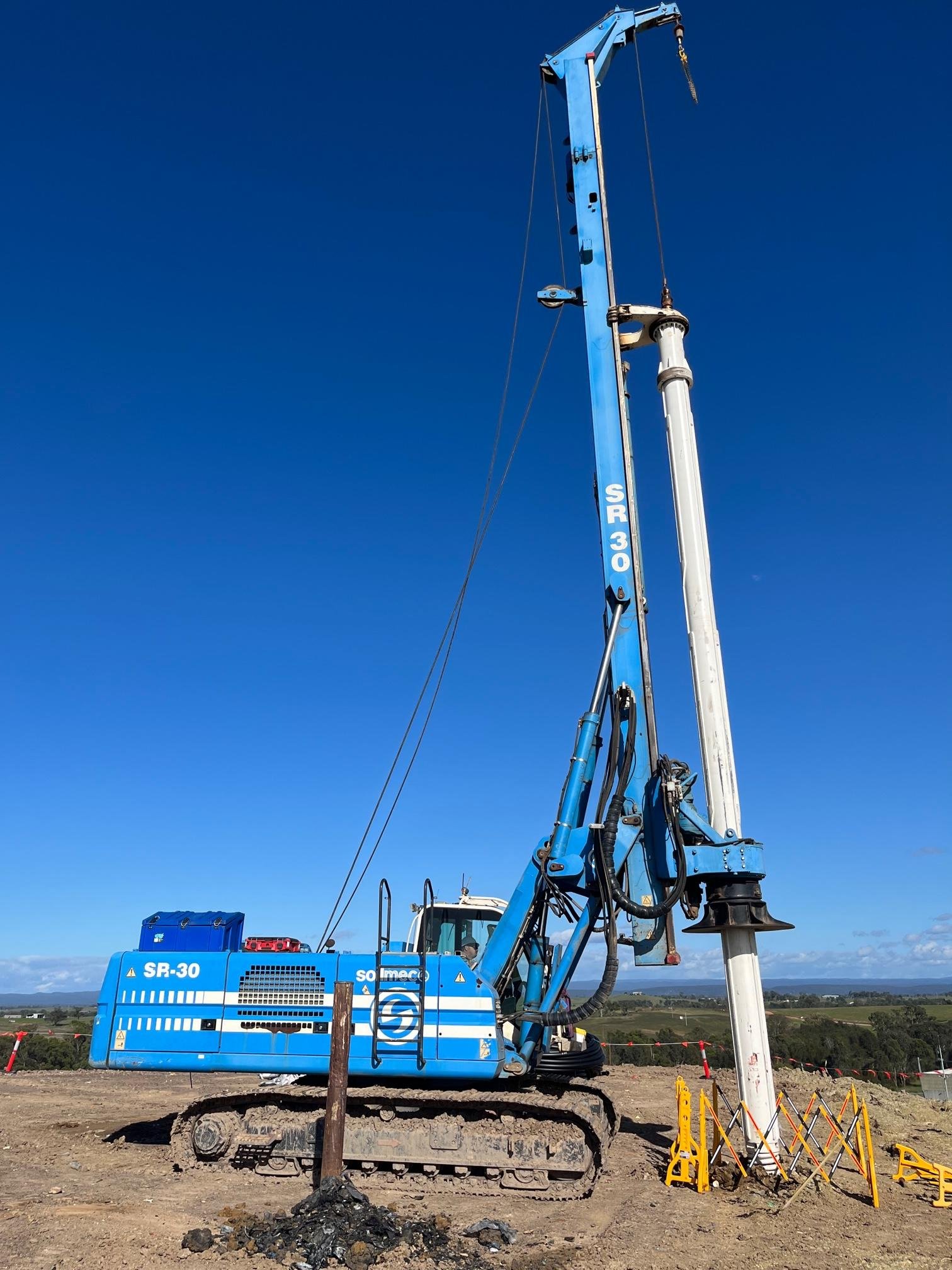A large blue drilling machine with the model number SR 30 is working at a construction site in a rural area under a clear blue sky. The machine has tracks, a tall vertical arm, and a white drilling component at the top. Yellow safety barriers and equipment are visible in the foreground.