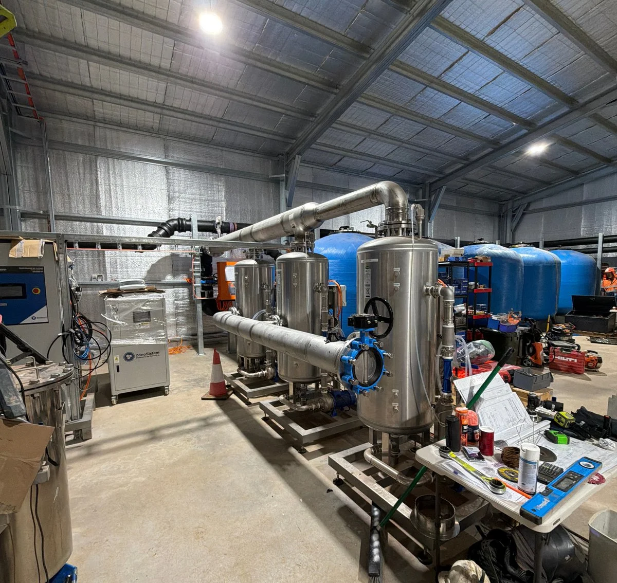 Industrial equipment setup with stainless steel tanks, blue water tanks, pipes, tools, and parts inside a warehouse.