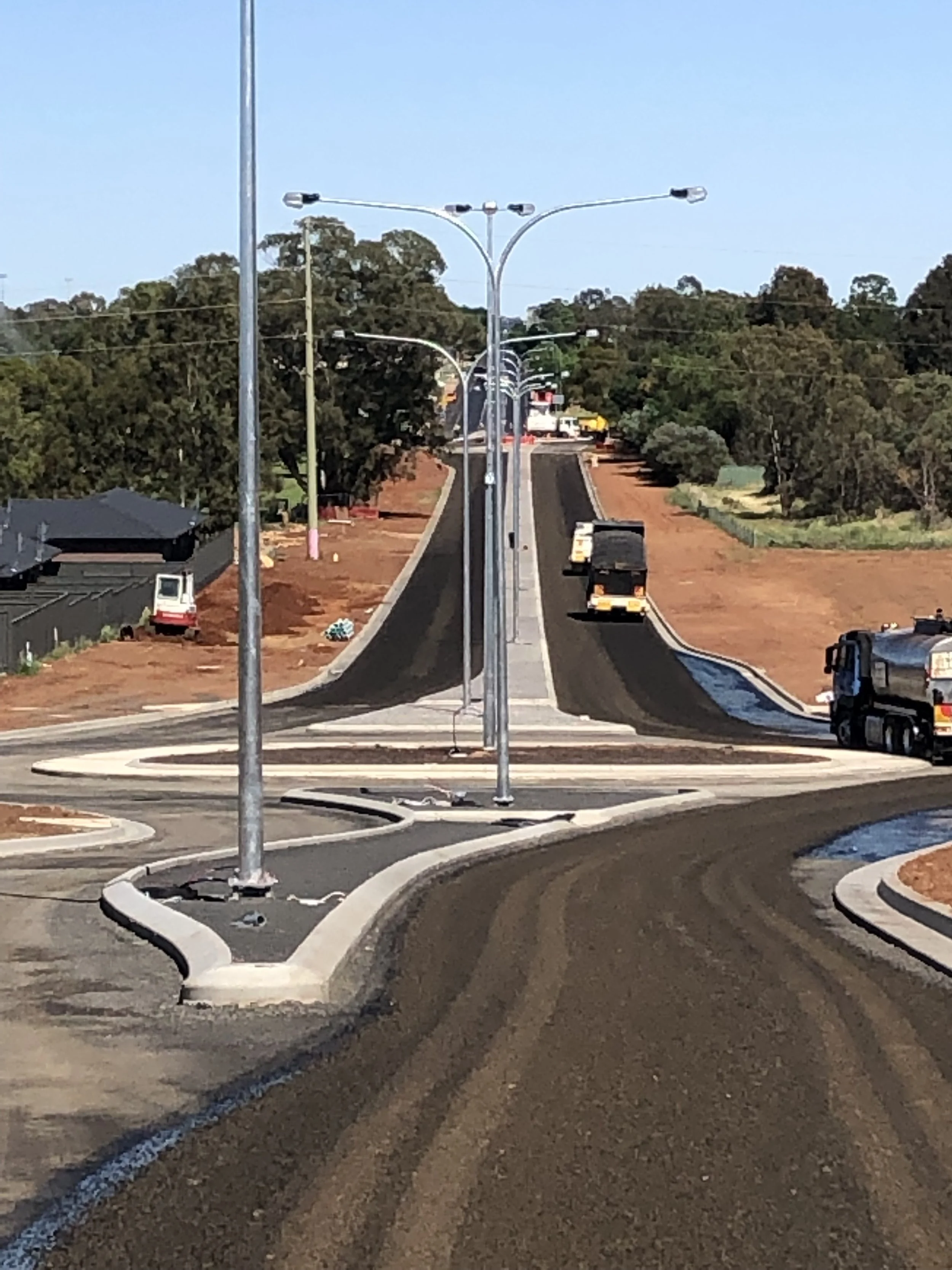 Photo of a newly constructed road with streetlights, surrounded by trees and some construction vehicles, during daytime.