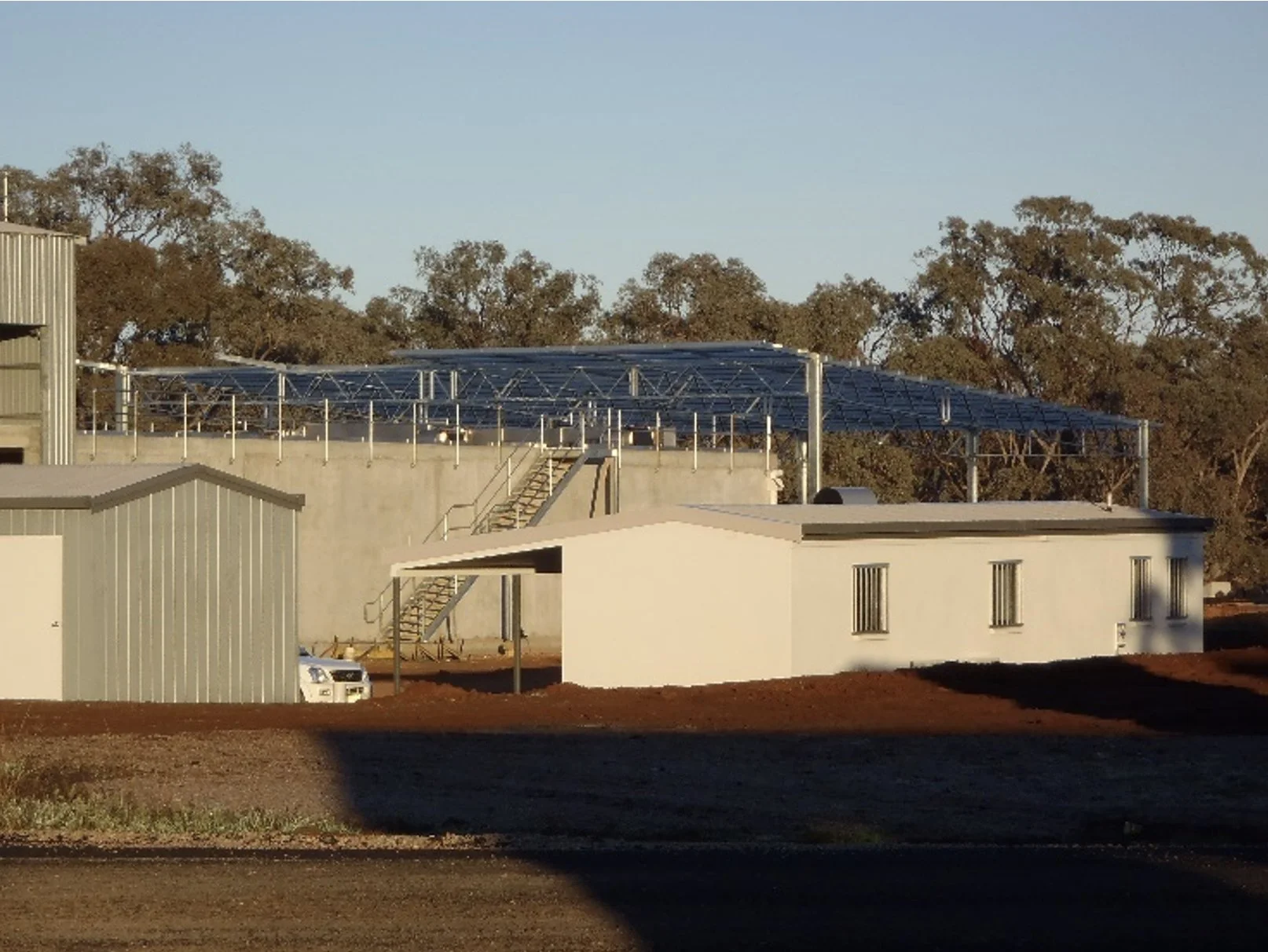 Building under construction with a large solar panel array on top, adjacent to several smaller structures, with trees in the background under a clear sky.