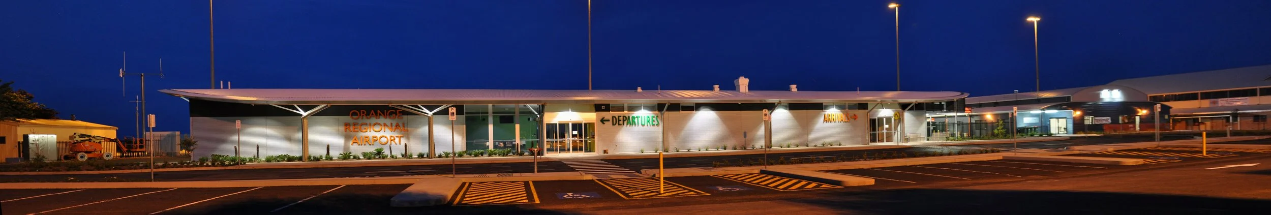 Night view of Orange Regional Airport with illuminated signs for departures and arrivals, empty parking lot in foreground.