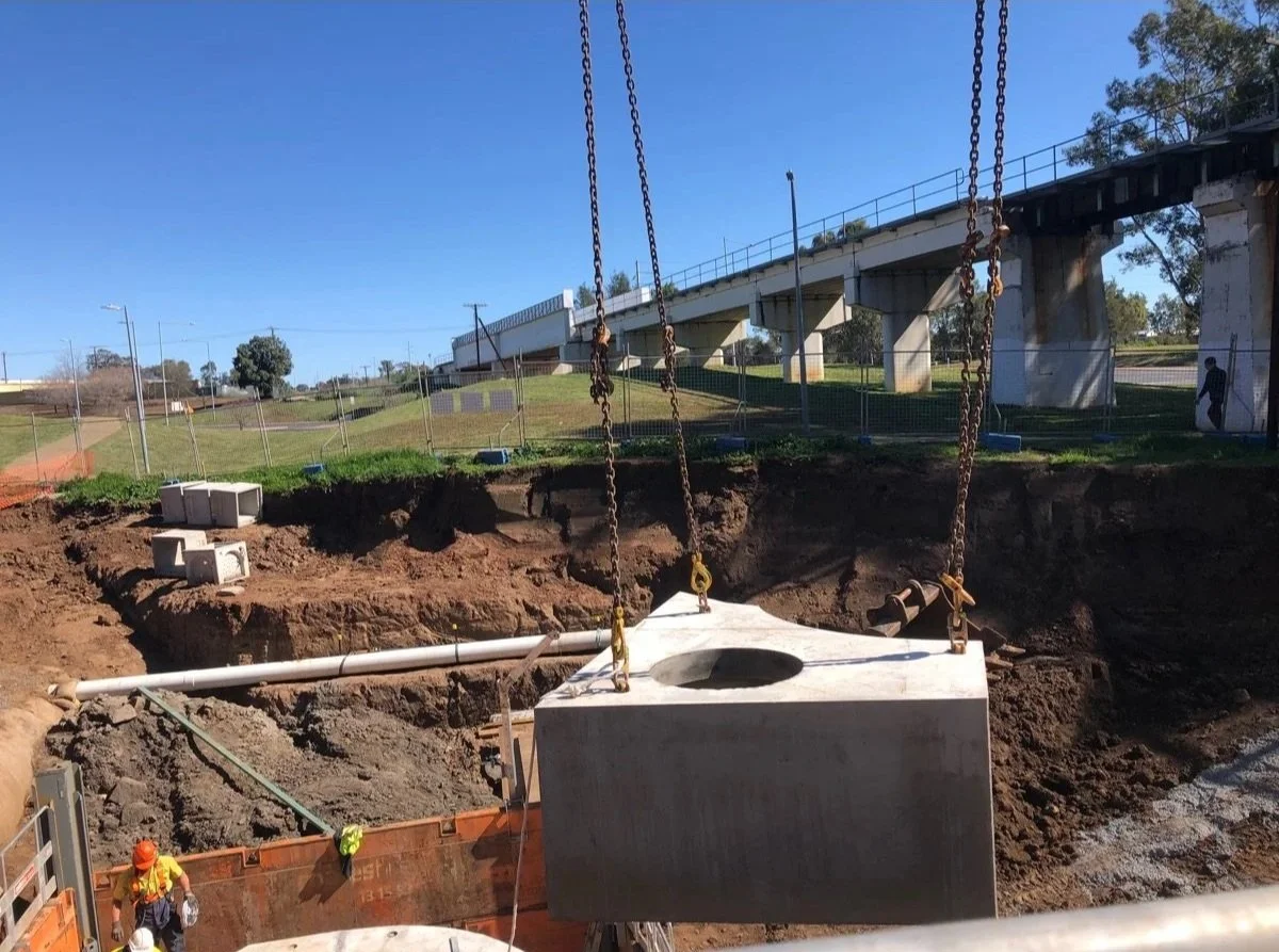 Construction site where a large concrete drainage box is being lowered into an excavation area with construction workers in safety gear and a pipe nearby, with a bridge overhead and a fence in the background.