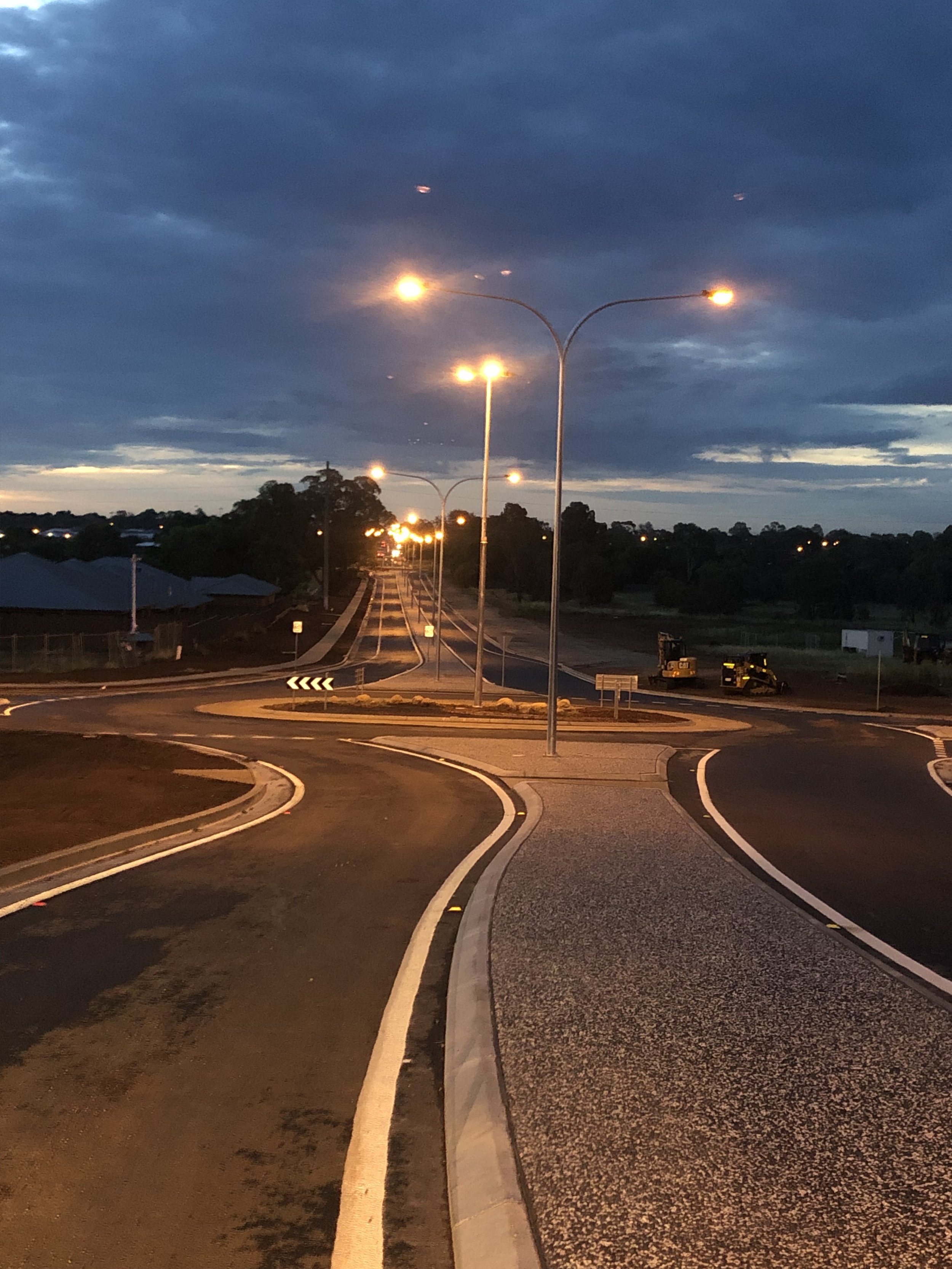 A newly paved roundabout with multiple streetlights illuminating the dark evening sky, leading into a residential area with houses and trees.