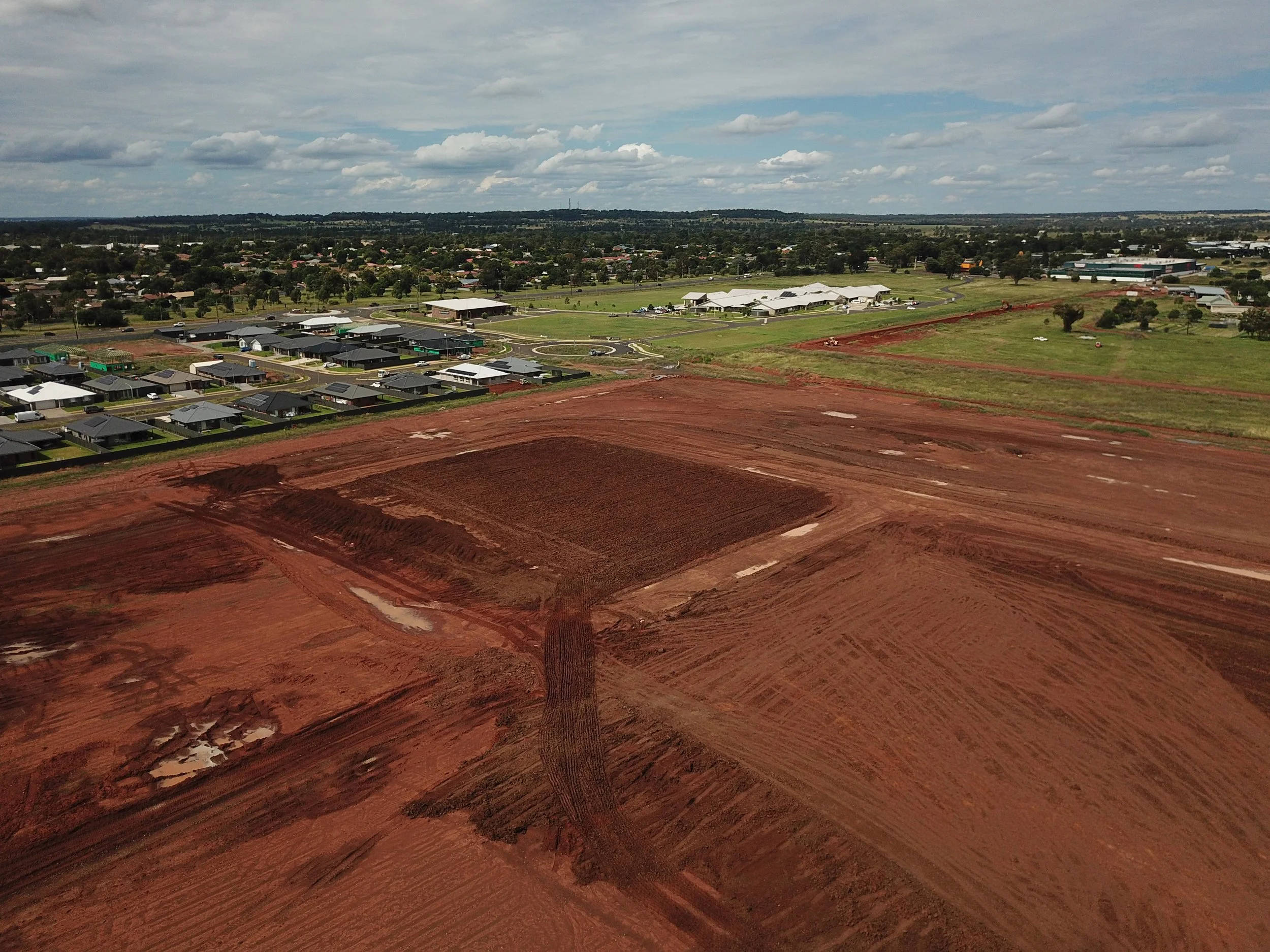 Aerial view of a suburban area with houses, roads, and green spaces, with a construction site with red soil in the foreground.