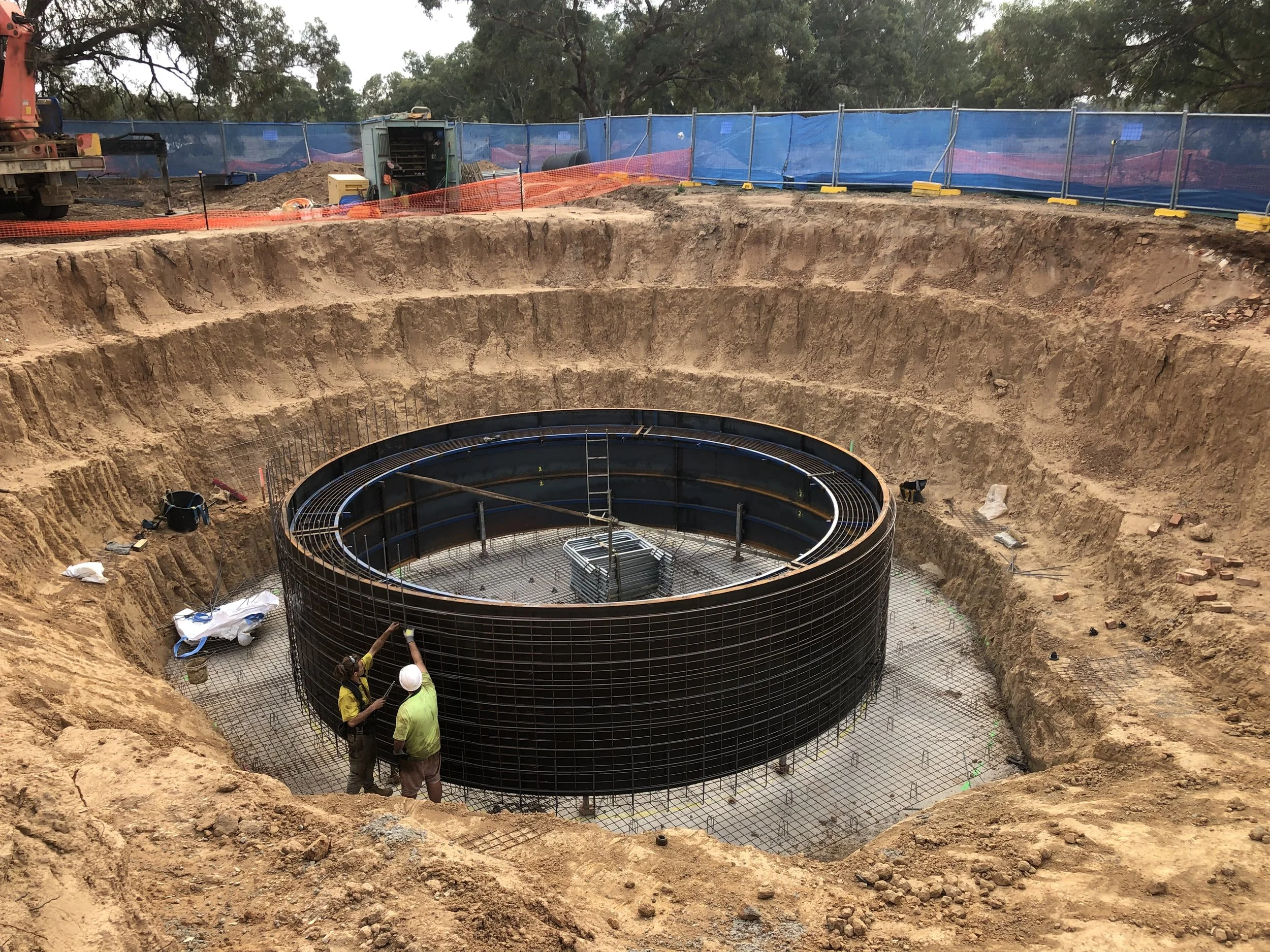 Construction site showing an excavated pit with a large circular steel reinforcement structure at the bottom, workers in safety gear, and equipment on the site with trees in the background.