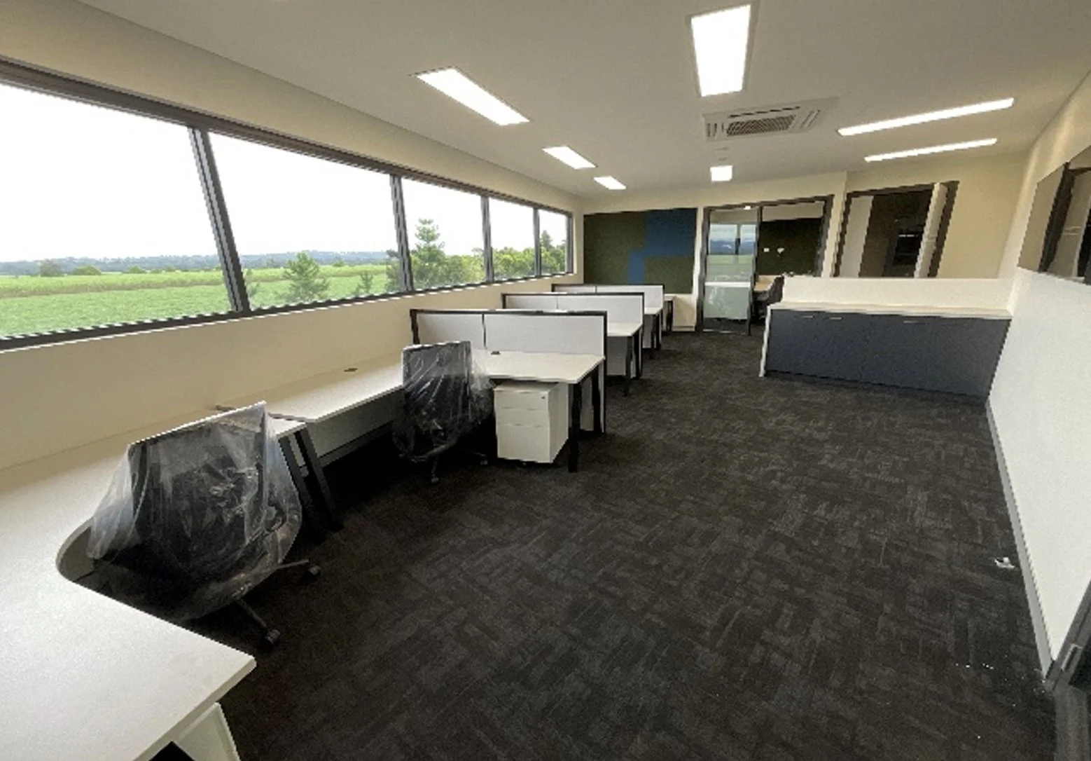 An empty office space with large windows overlooking a green landscape, white desks with black frames, some chairs covered in plastic, and a dark carpeted floor.