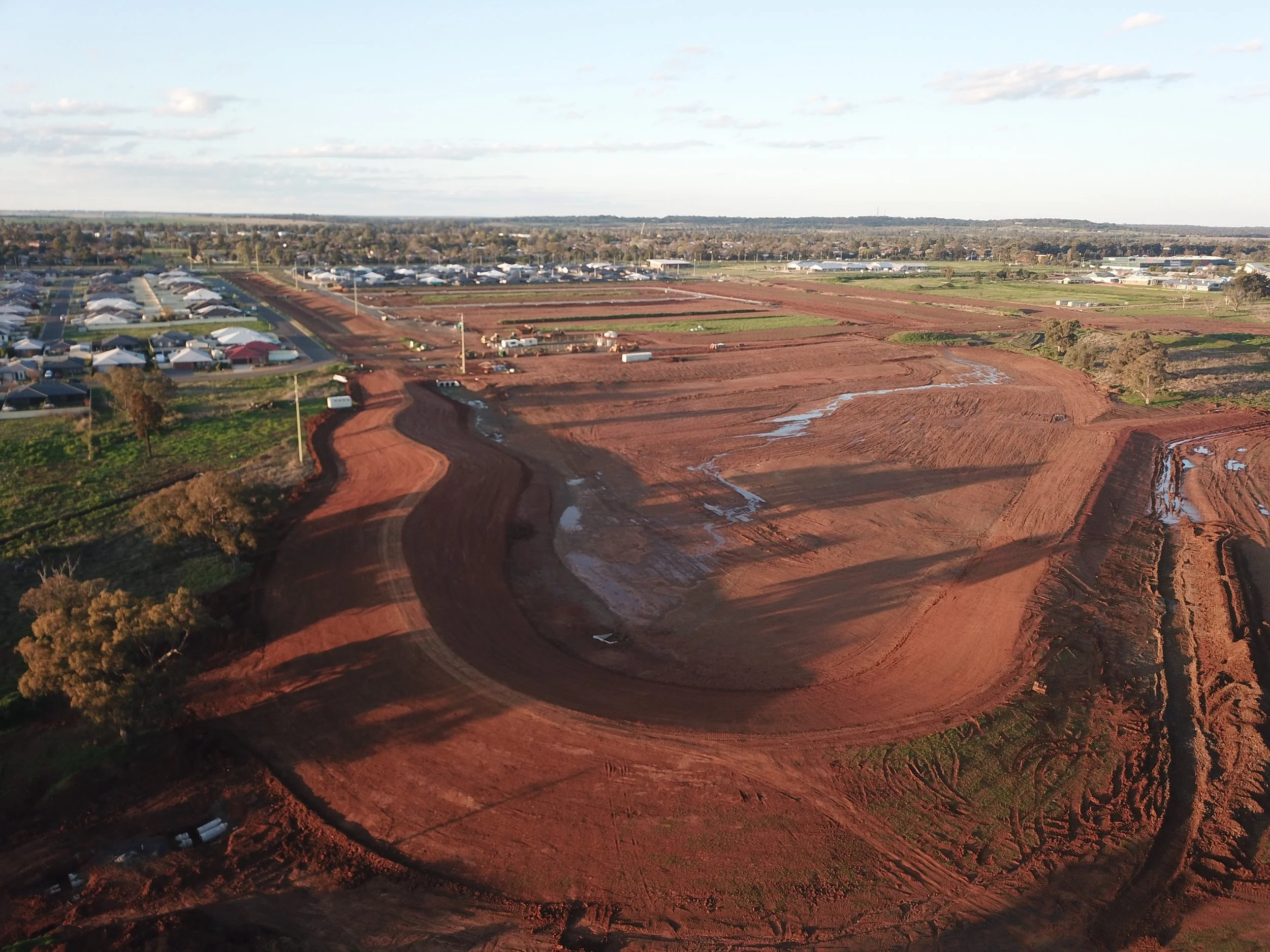 An aerial view of a construction site with red dirt, partially completed roads, and surrounding green vegetation, with residential houses in the background under a partly cloudy sky.