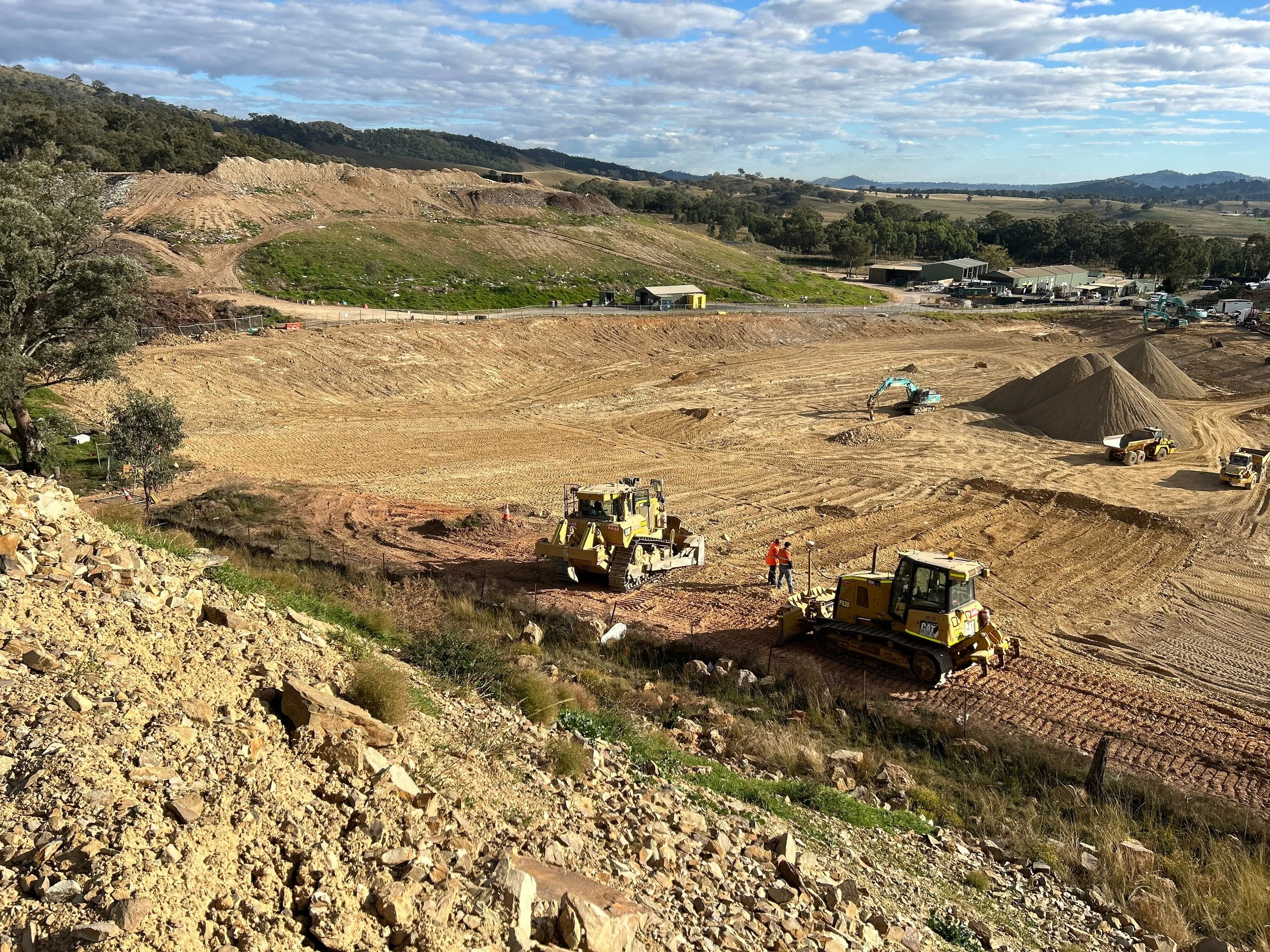 Construction site with heavy machinery and earth-moving equipment; piles of dirt and construction workers working on the site, with a background of green hills and a partly cloudy sky.