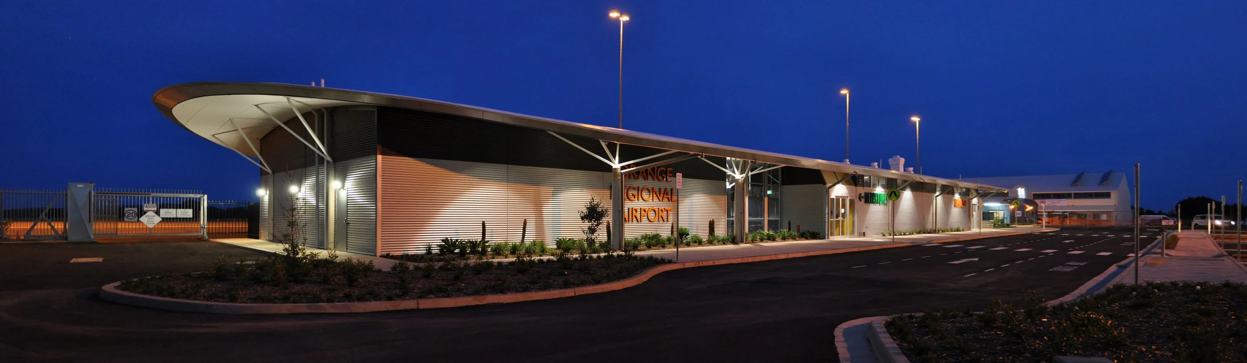 Night view of a modern international airport terminal with a curved roof, illuminated signs, and parking lot in the background.