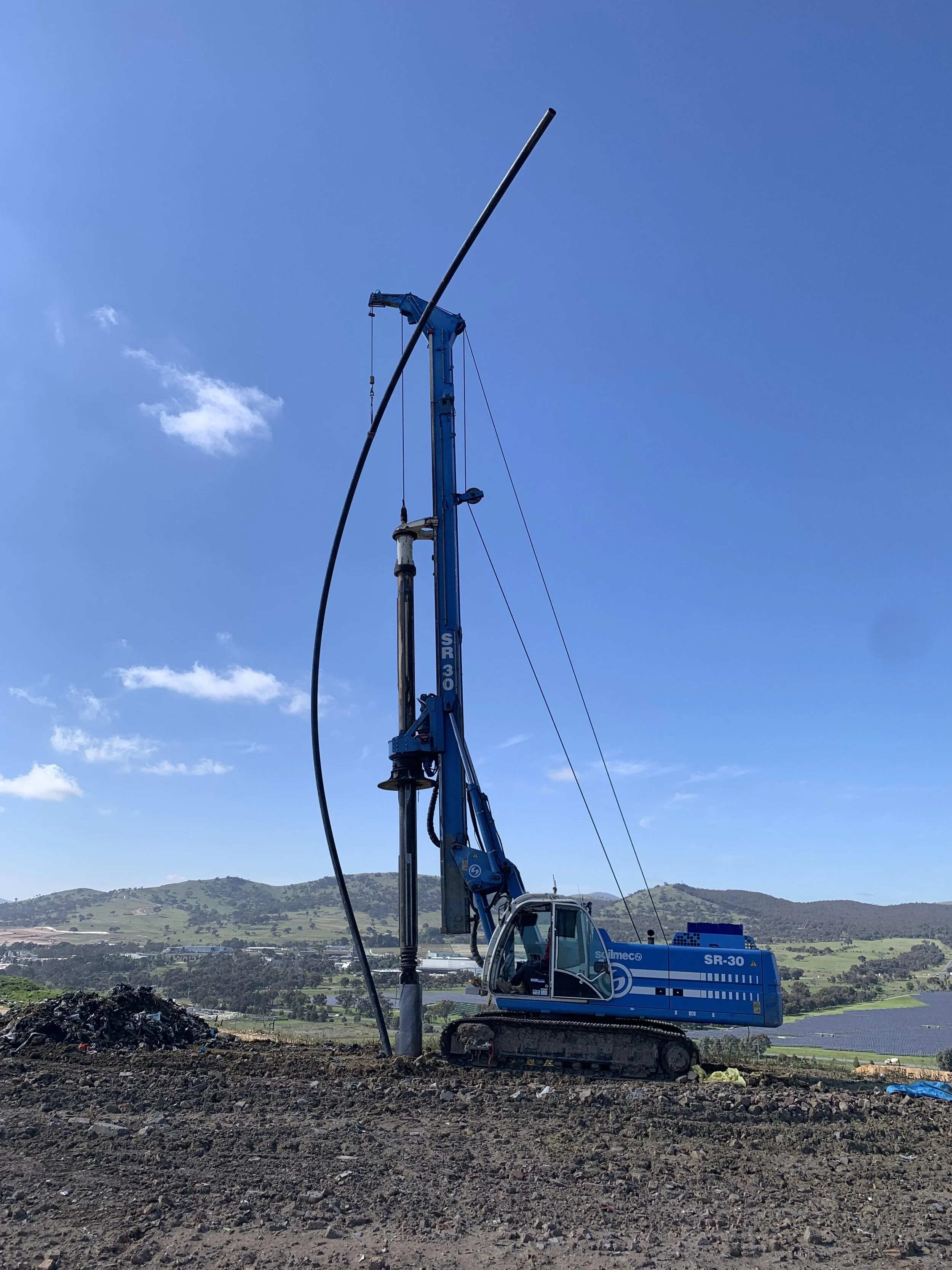 A blue construction excavator with a boom and a long pipe attached, working on a construction site with a backdrop of distant hills and a partly cloudy sky.
