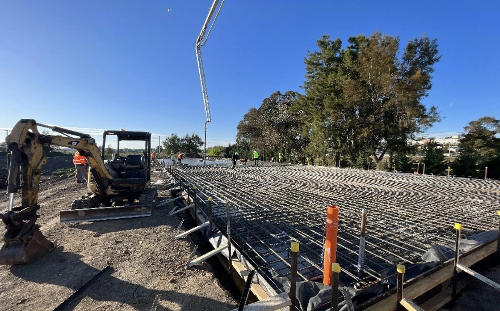 Construction site with rebar and a mini excavator, workers in the background, and a clear blue sky with a tree.