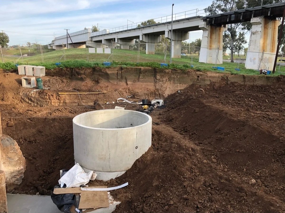 Construction site with a large concrete pipe on the ground, dirt excavations, and a bridge in the background.
