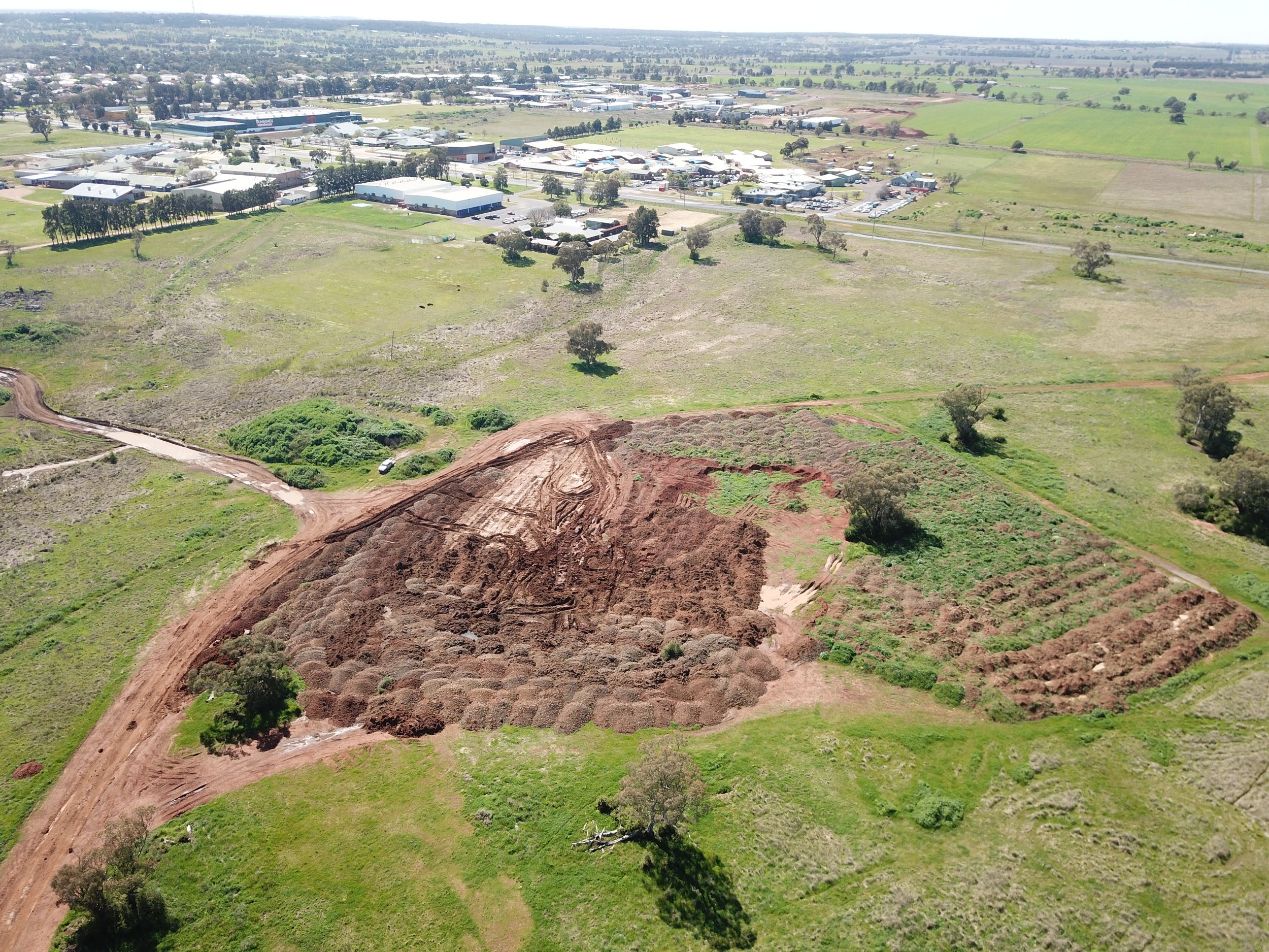 Aerial view of a construction site with large dirt mounds and surrounding green fields, with a small rural town or industrial area in the background.