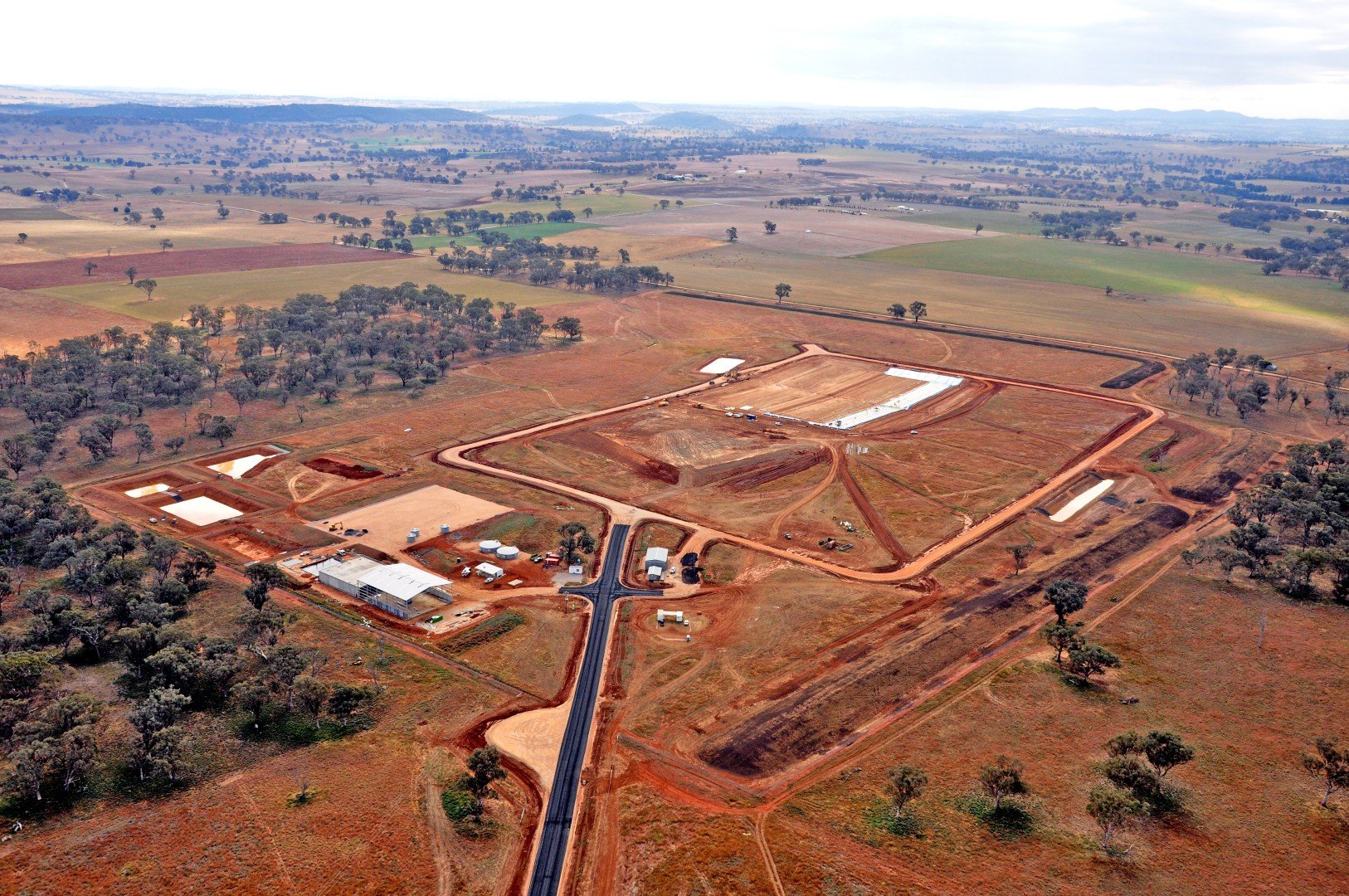 Aerial view of construction site with roads, foundation work, and some buildings in a rural landscape with trees and fields.