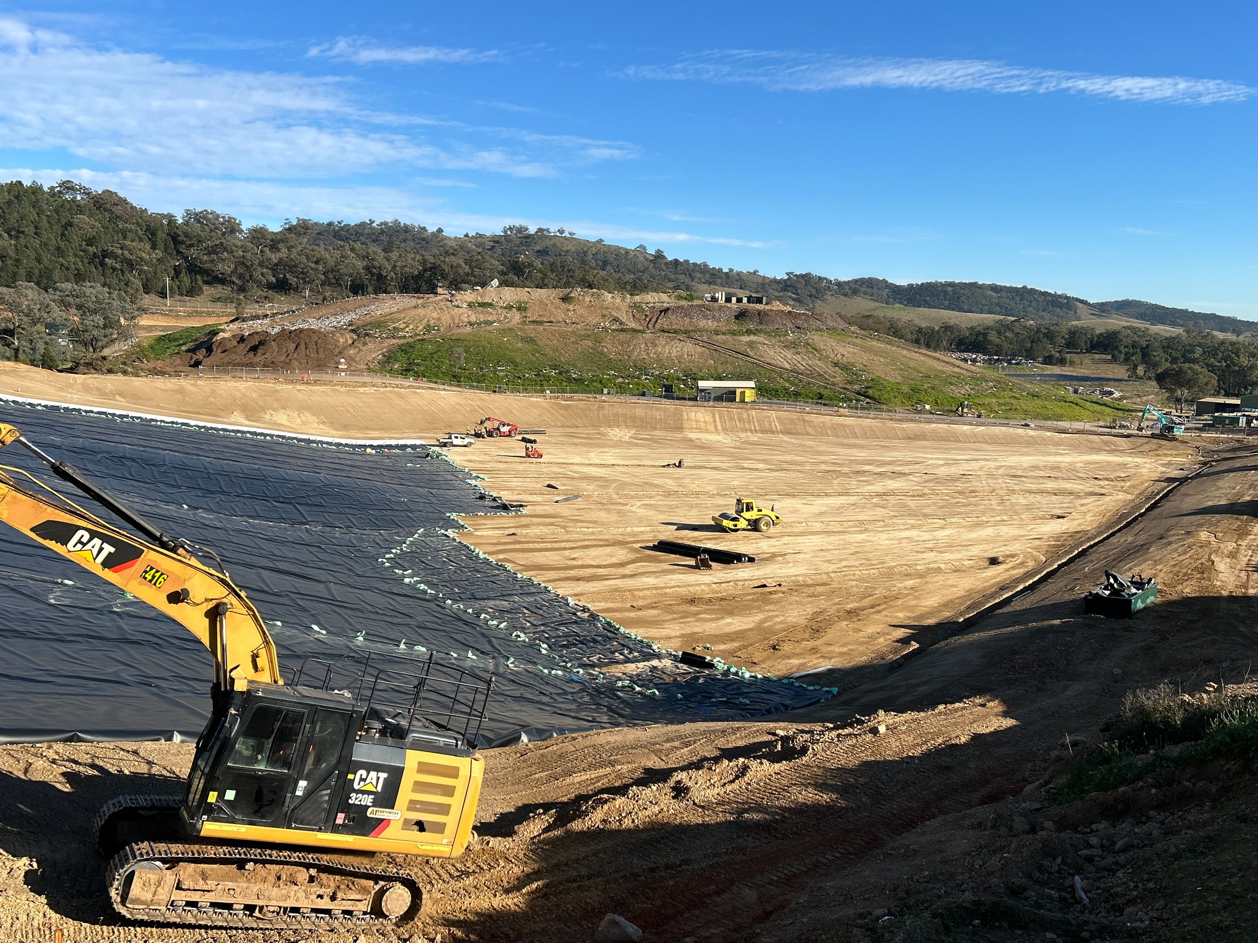 Construction site with a large area of dirt being prepared, multiple construction vehicles including excavators and trucks, and a hill in the background under a blue sky with some clouds.