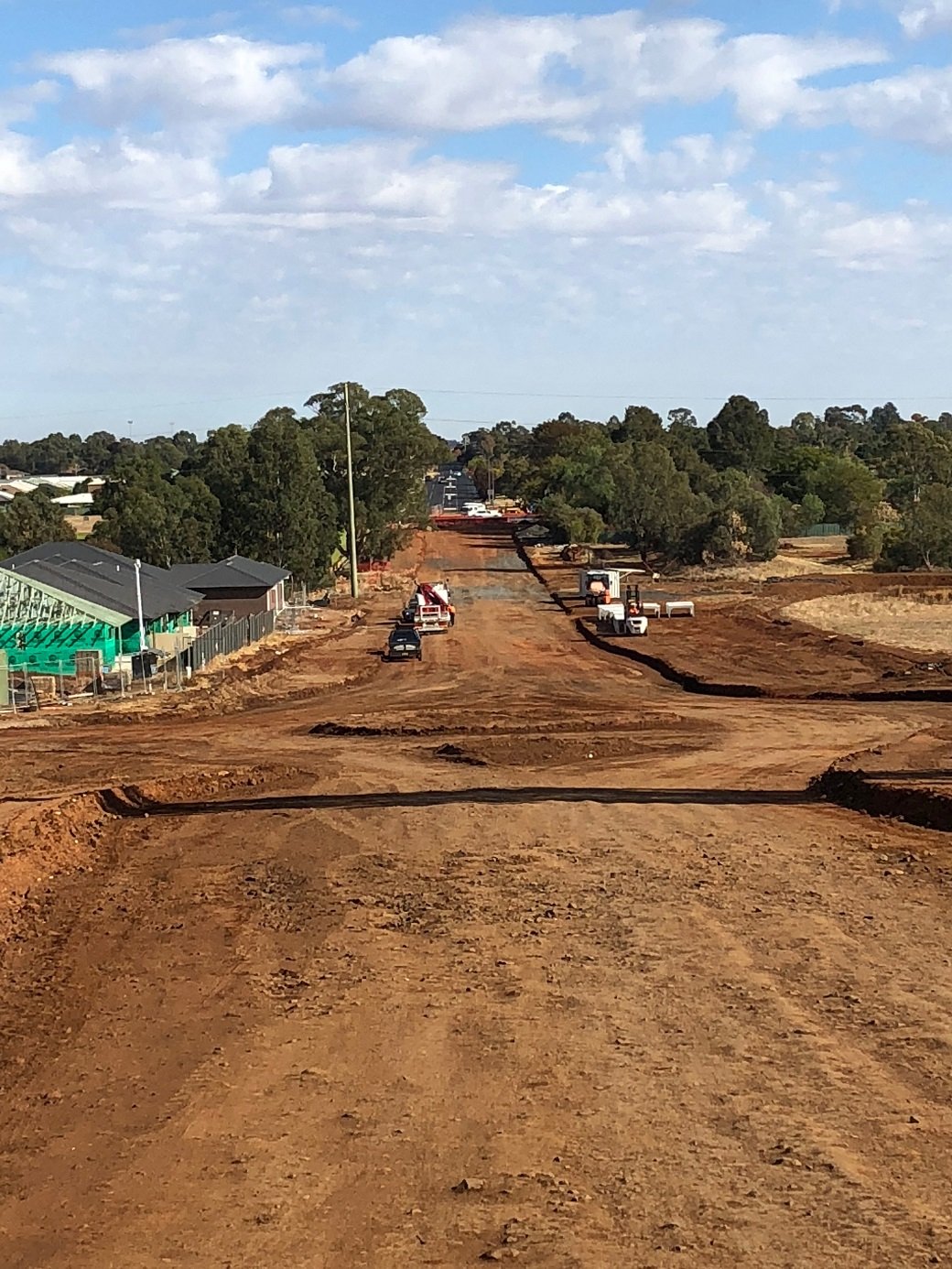Construction site with dirt road, construction vehicles, and trees in the background under partly cloudy sky.
