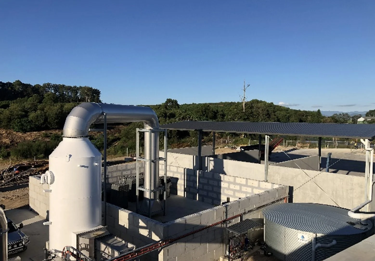 Construction site with solar panel roof, white industrial piping, and heat exchange equipment, set against a backdrop of trees and a clear blue sky.