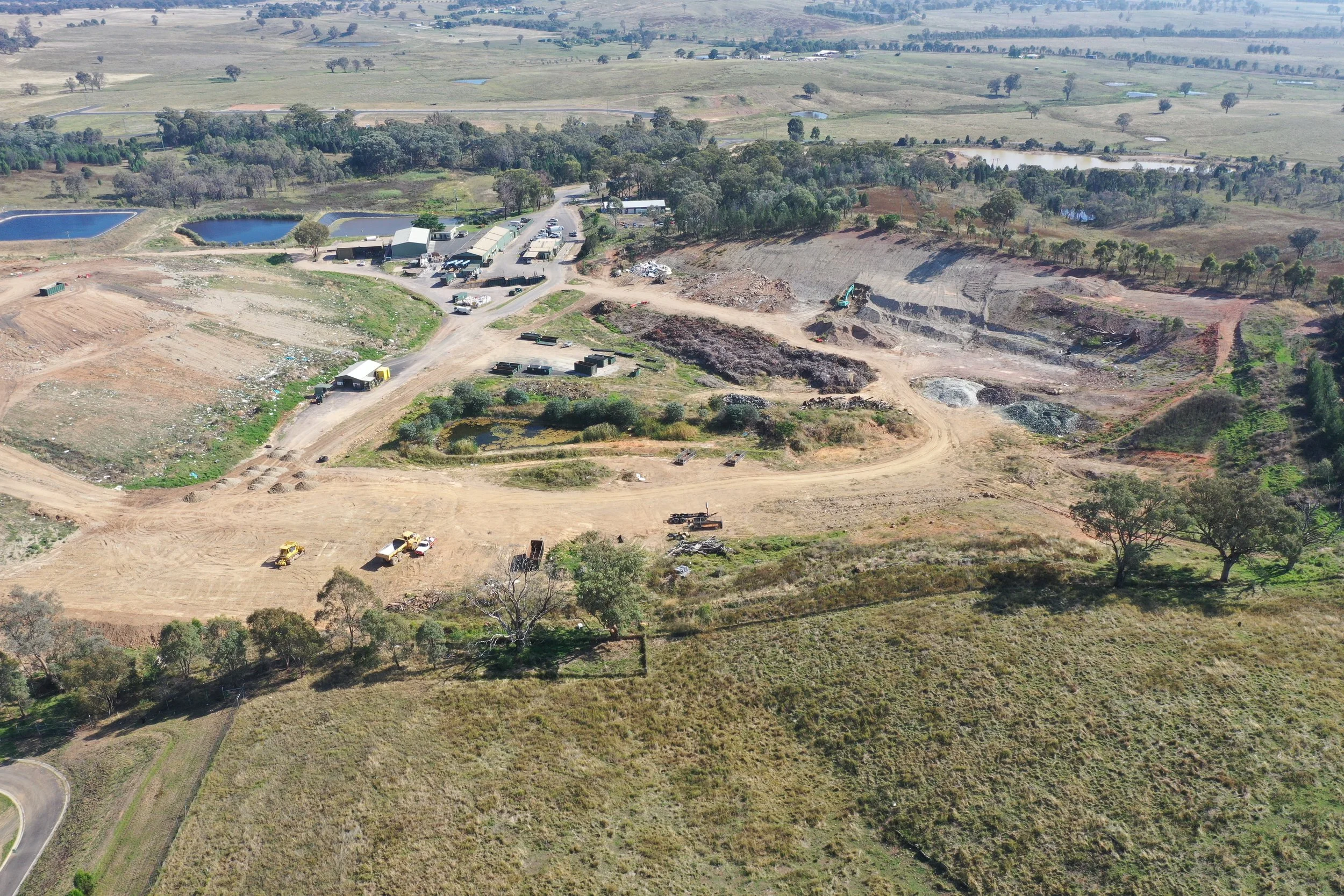 Aerial view of a construction site in a rural area with dirt roads, construction equipment, storage containers, and partially built structures surrounded by green landscape and small ponds.