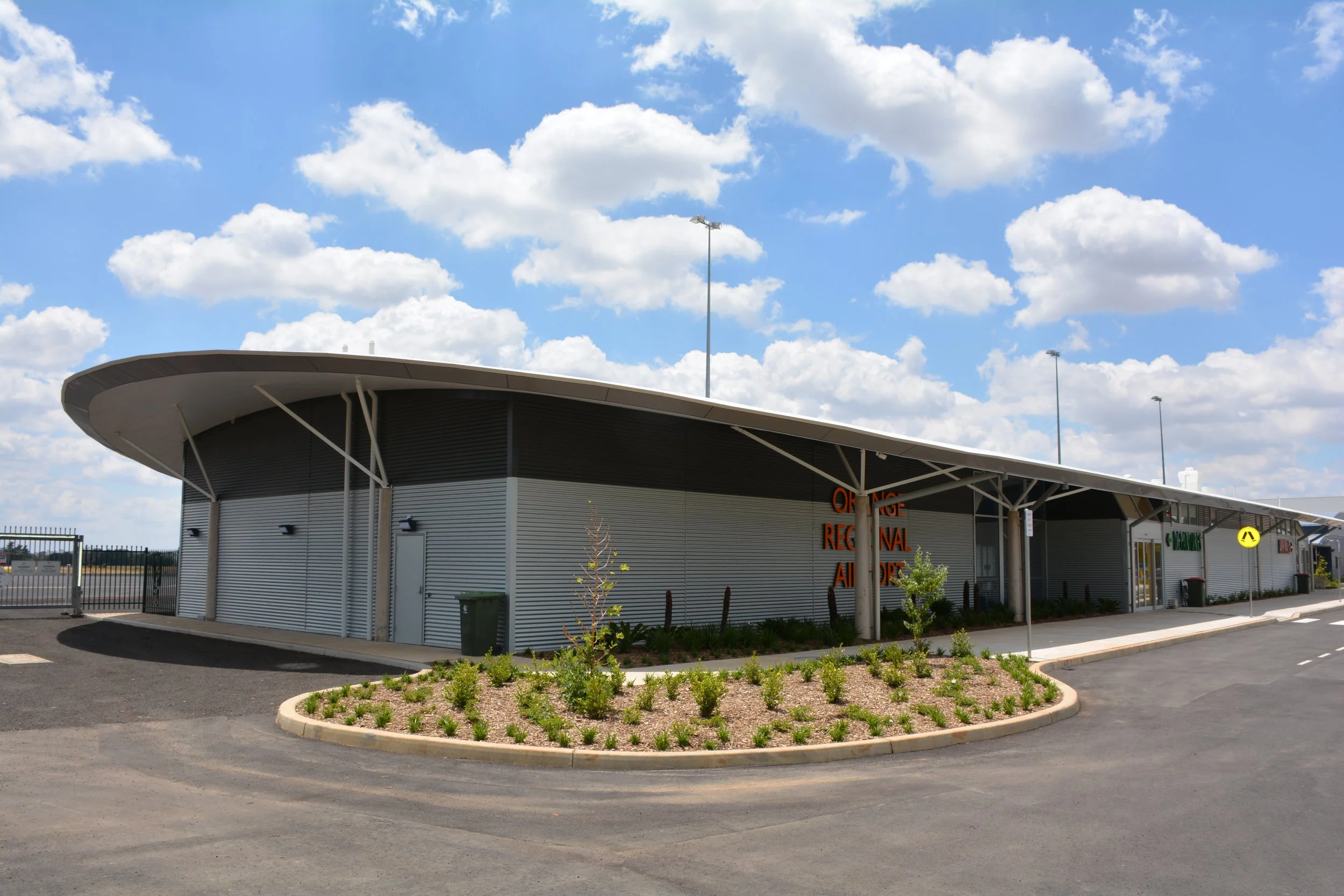 Modern airport terminal building with a curved roof, surrounded by a parking lot and landscaped area, under a partly cloudy sky.