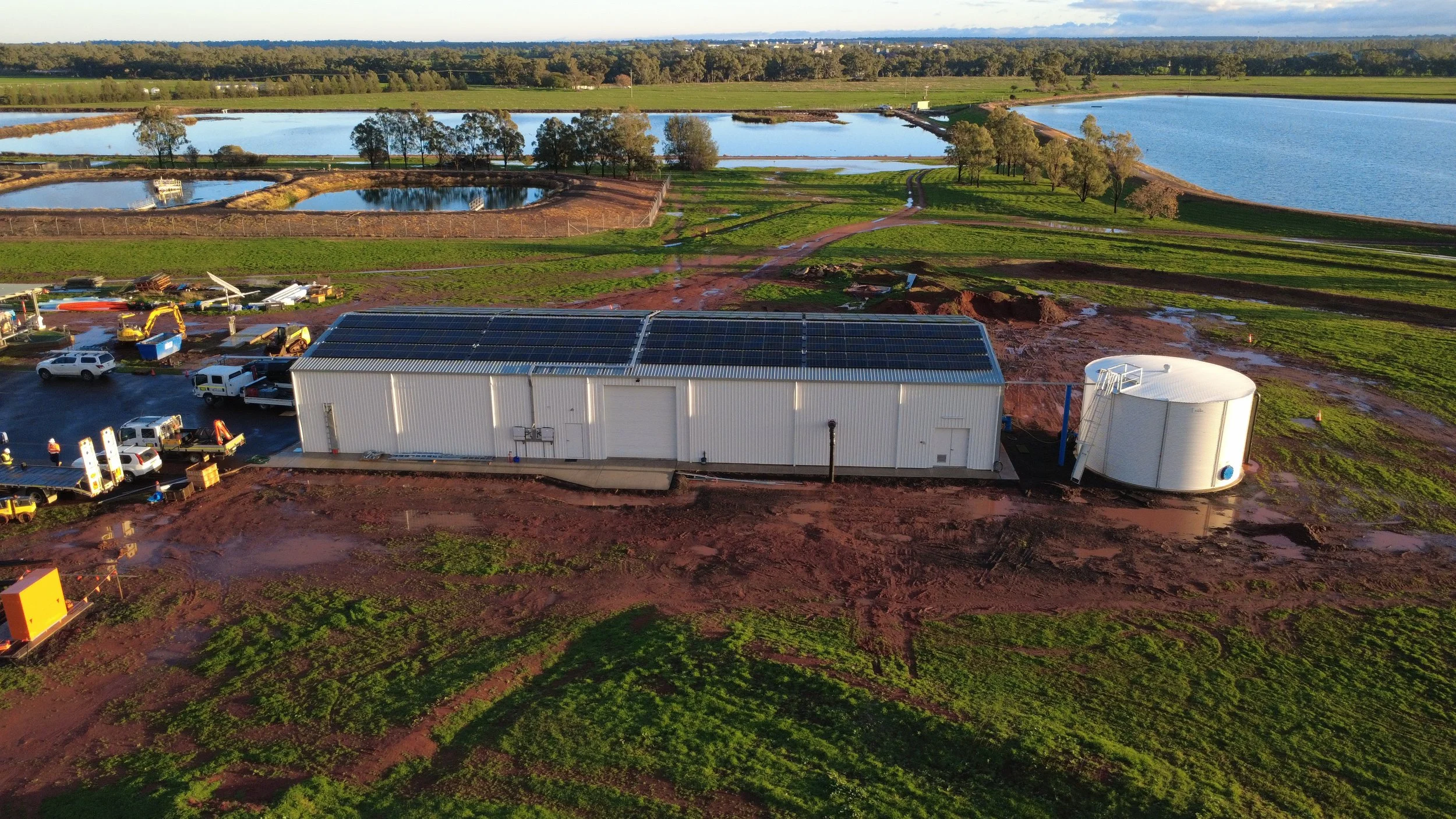 Aerial view of a water treatment facility with large tanks, green fields, and patches of red soil, surrounded by water bodies and trees.
