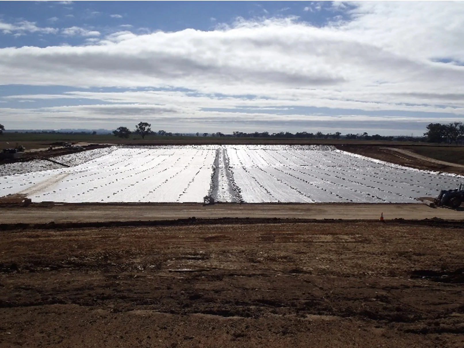 A large agricultural field covered with white plastic sheeting for crop planting, with a dirt road in the foreground and a partly cloudy sky overhead.