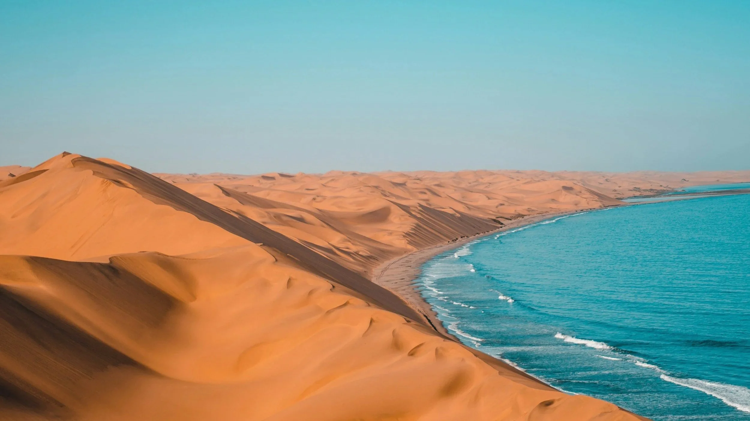 Sand dunes alongside a turquoise ocean with gentle waves, under a light blue sky.
