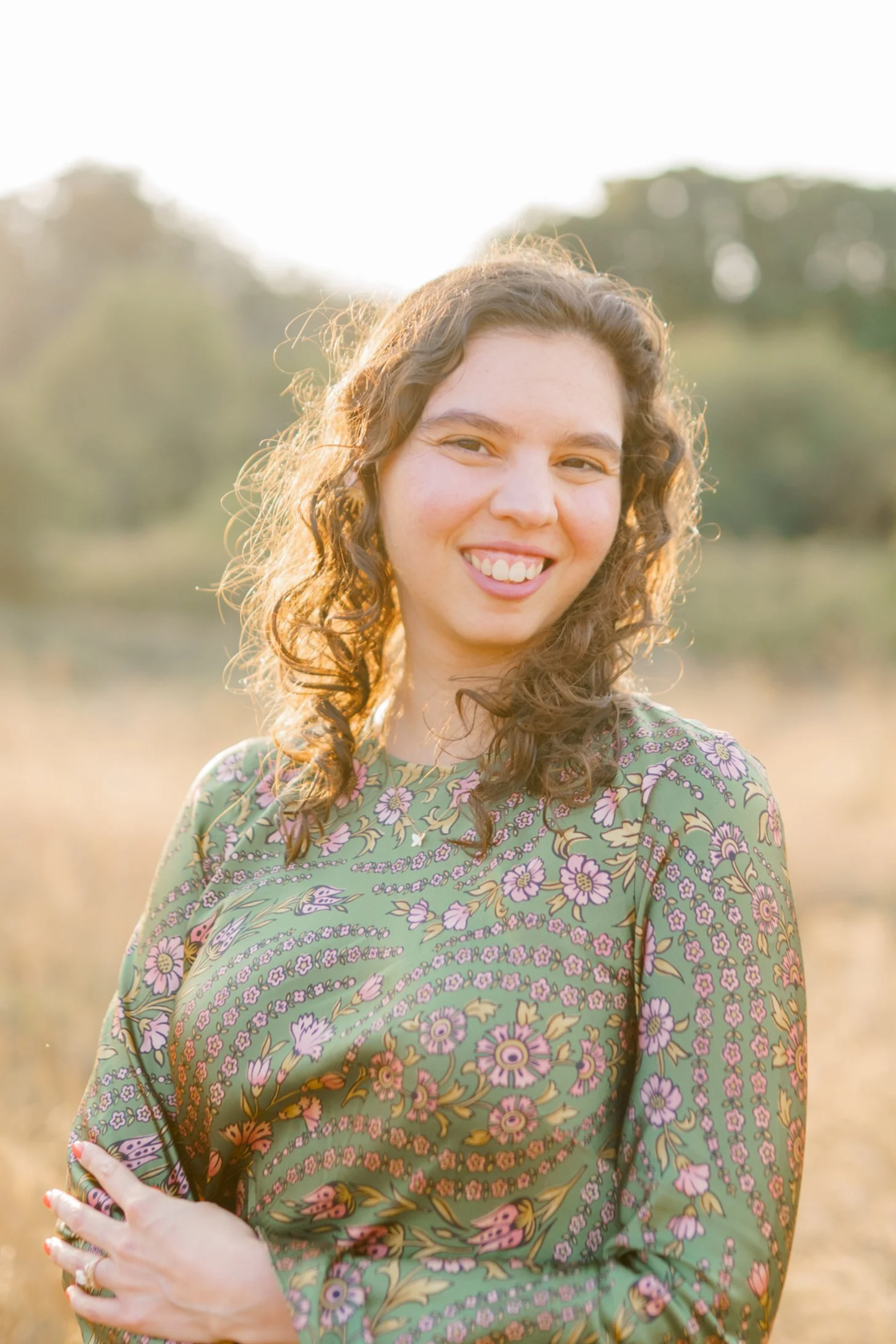 The mediator ready to help you, smiling with curly hair wearing a patterned dress standing outdoors in a field during sunset.