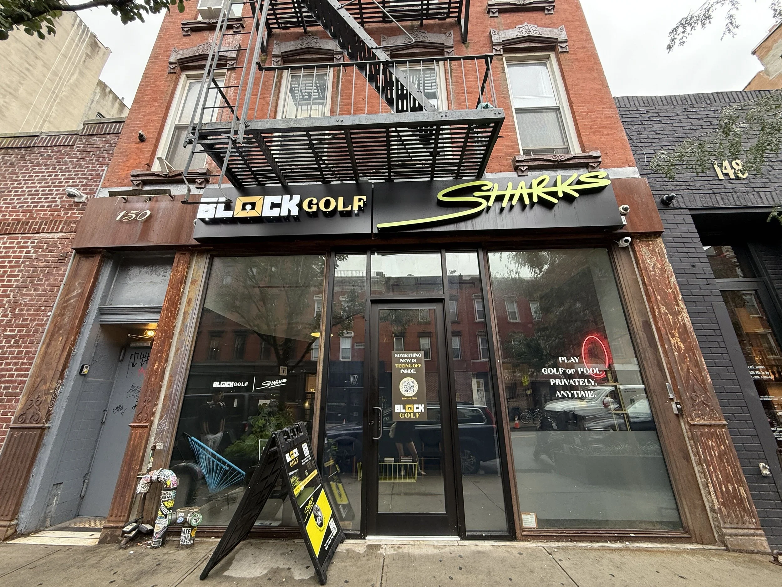 Storefront with signs for Block Golf and Sharks, with a glass door and window at street level, featuring a sidewalk with a skateboard and a shopping cart outside.