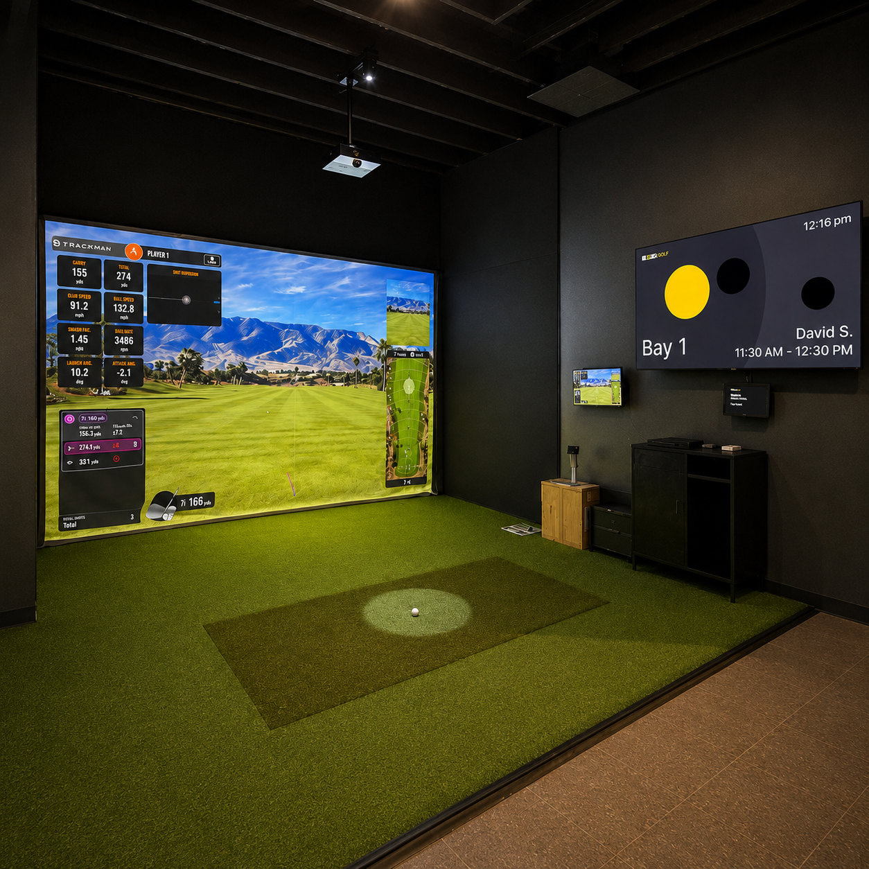 Indoor golf simulation room with a large screen displaying a virtual golf course, smaller screens on the wall showing golf information and booking details, a golf tee on a small mat, and artificial grass covering the floor.