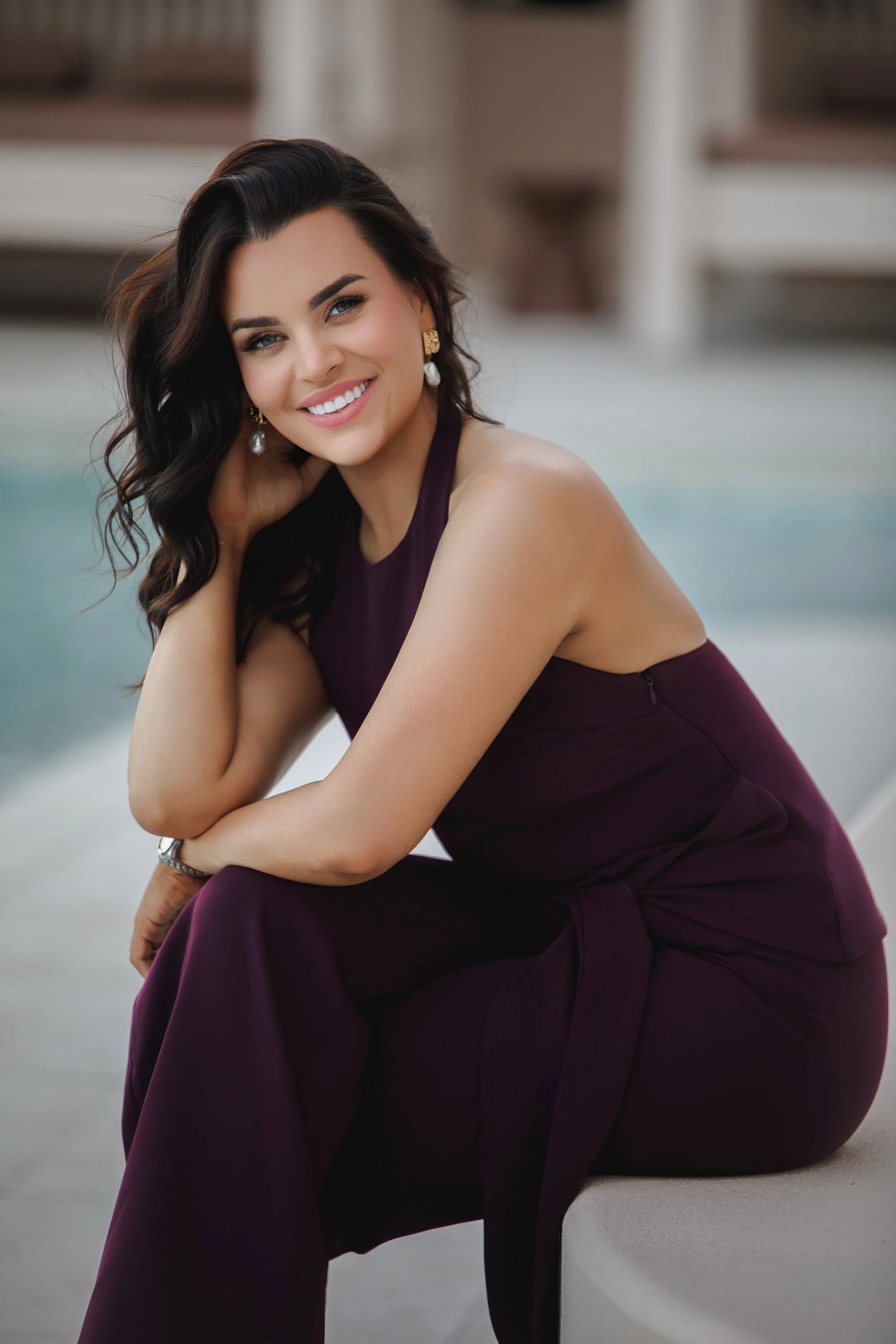 A smiling woman with dark wavy hair, wearing pearl earrings and a dark purple sleeveless dress, sitting by a pool or water feature outdoors.
