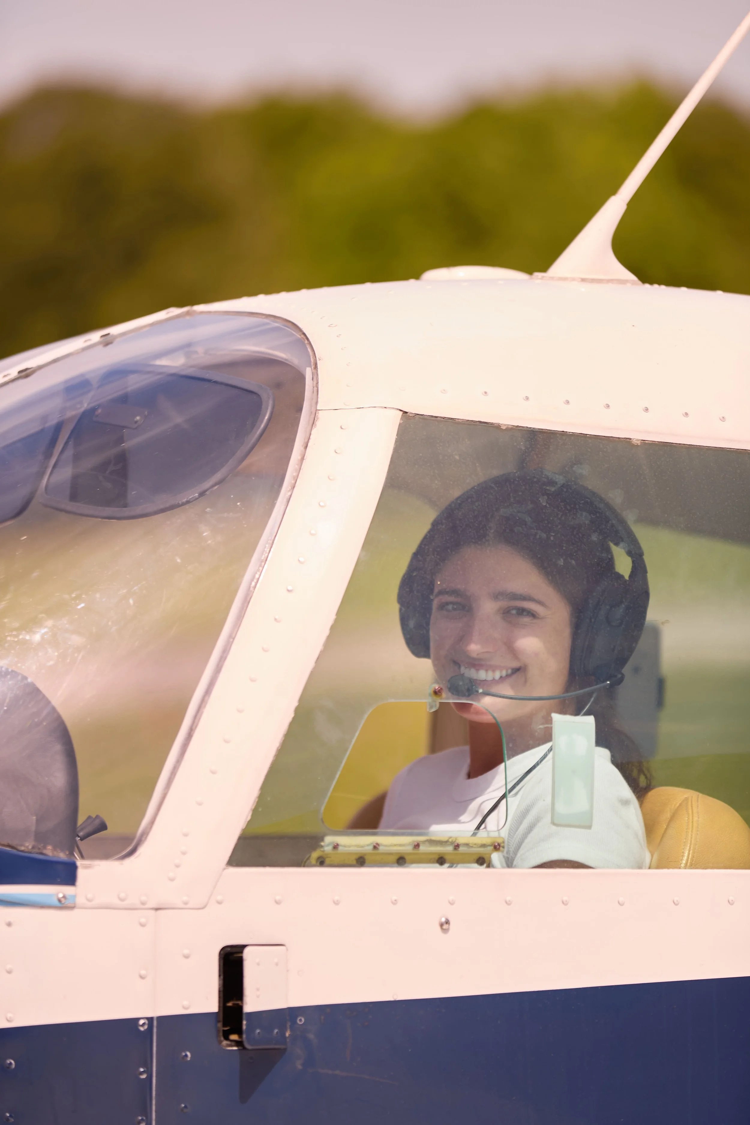 A smiling woman wearing headphones sitting inside the cockpit of an aircraft, visible through the cockpit window.