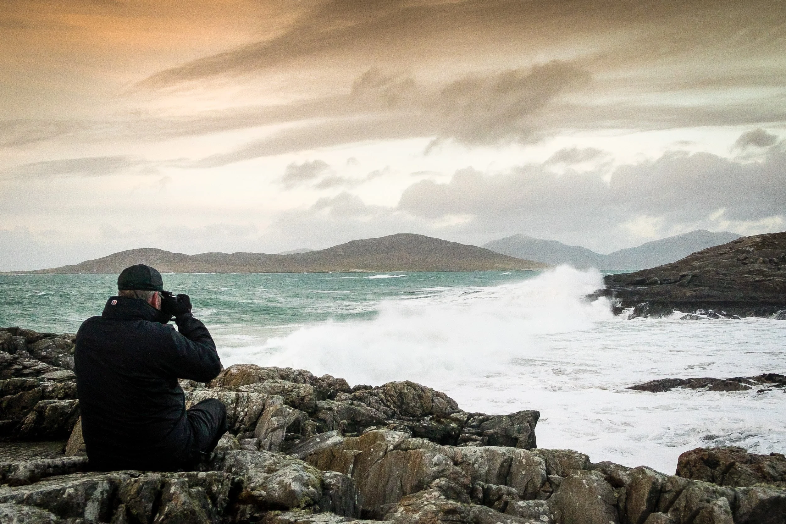 A person sitting on rocks near the shore of a body of water, taking a photo of the ocean with mountains and cloudy sky in the background | Steven Marshall Photography