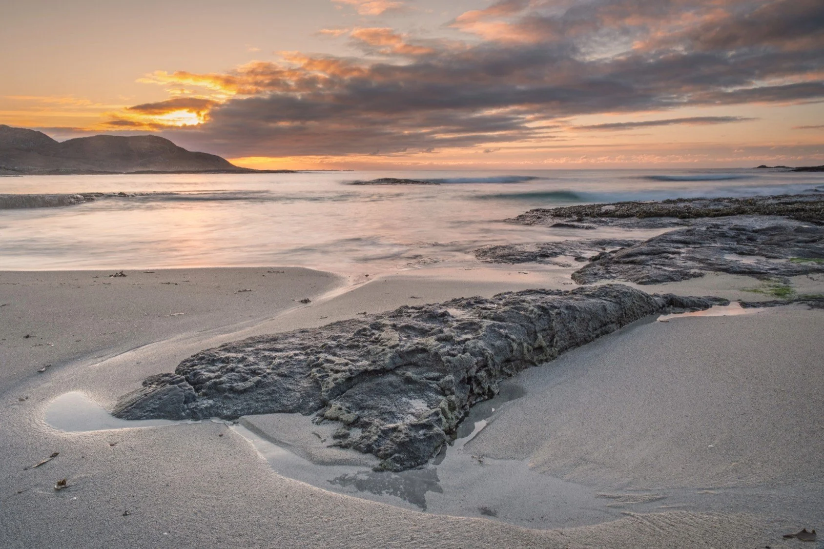 The sun setting behind the hills on the west side of Sanna Bay, with waves lapping on the sandy beach and rocks and sunset colour clouds in the sky | Ardnamurchan Scotland | Steven Marshall Photography