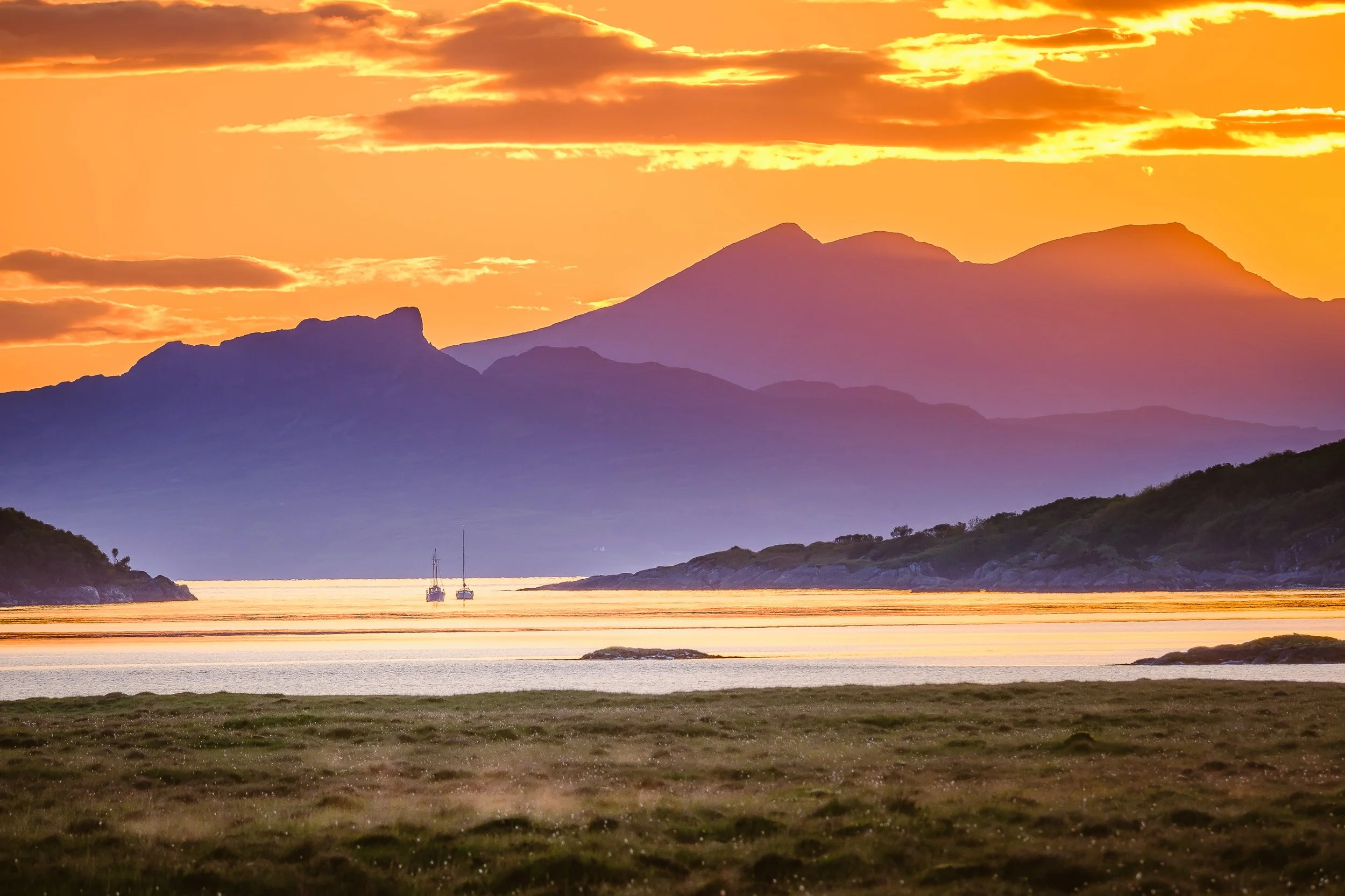 A scenic sunset over the mountain range of the Isle of Rum with a body of water in the foreground and two sailboats floating on it | | Steven Marshall Photography