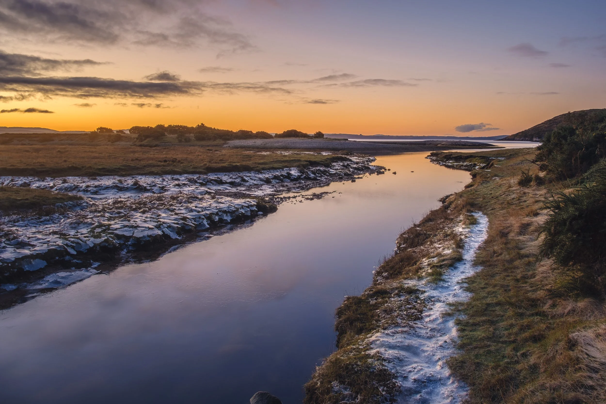 A winter morning sunrise over a river with grassy banks covered in thick frost, with hills and clouds in the background | Steven Marshall Photography