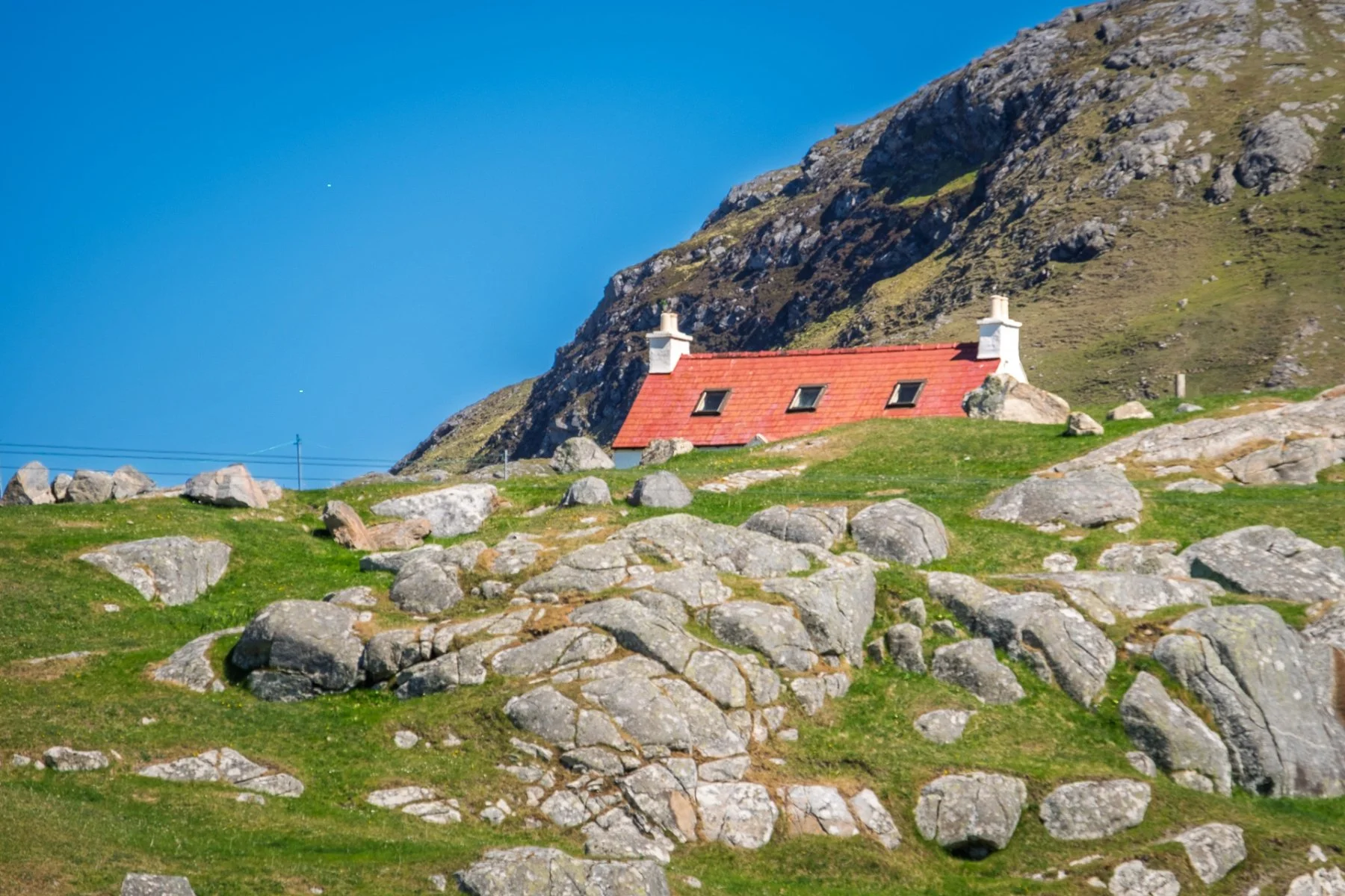 The red roof of a white croft house in Am Baile on Eriskay peaking above a rock and grass covered foreground | Outer Hebrides Scotland | Steven Marshall Photography