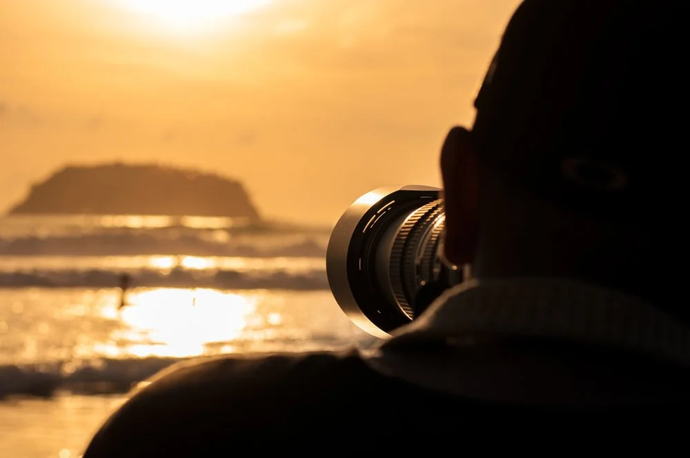 A person using a camera to photograph a sunset over the ocean, with a small island silhouette in the background | Steven Marshall Photography