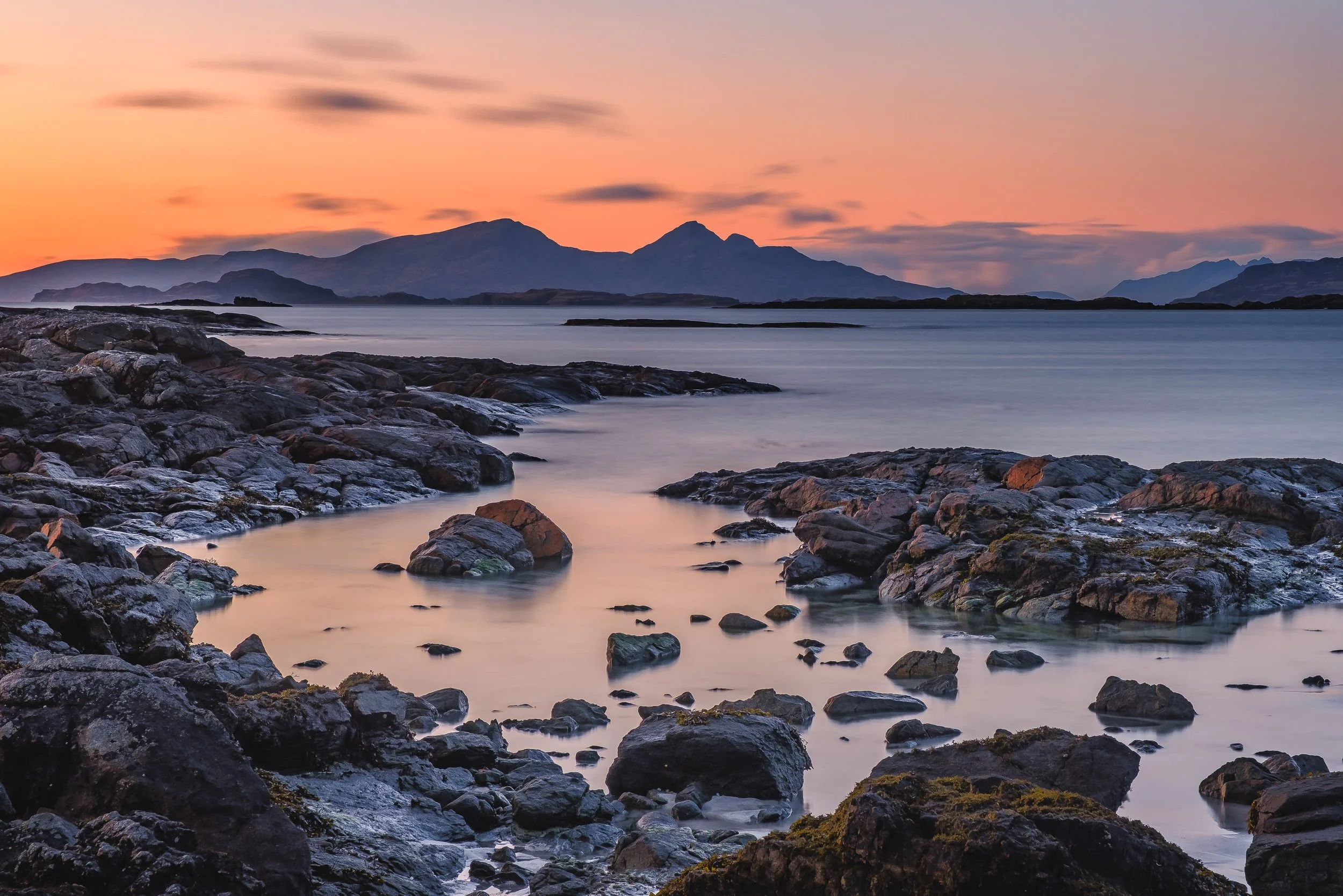 Rocks along the shoreline at Portuairk, Ardnamurchan with calm water and mountains of the Isle of Rum in the background during sunset with a pinkish sky | Steven Marshall Photography