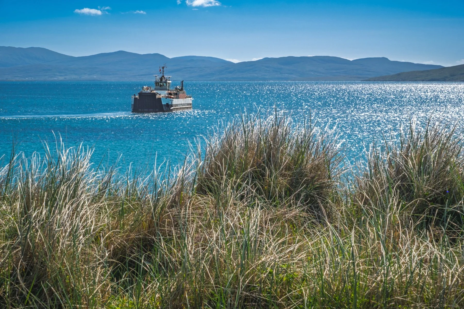 CALMAC’s MV Loch Alainn leaving the ferry terminal on Eriskay and heading towards the Isle of Barra on the horizon. Marram grass in the foreground, blue sea and clear blue sky | Outer Hebrides Scotland | Steven Marshall Photography
